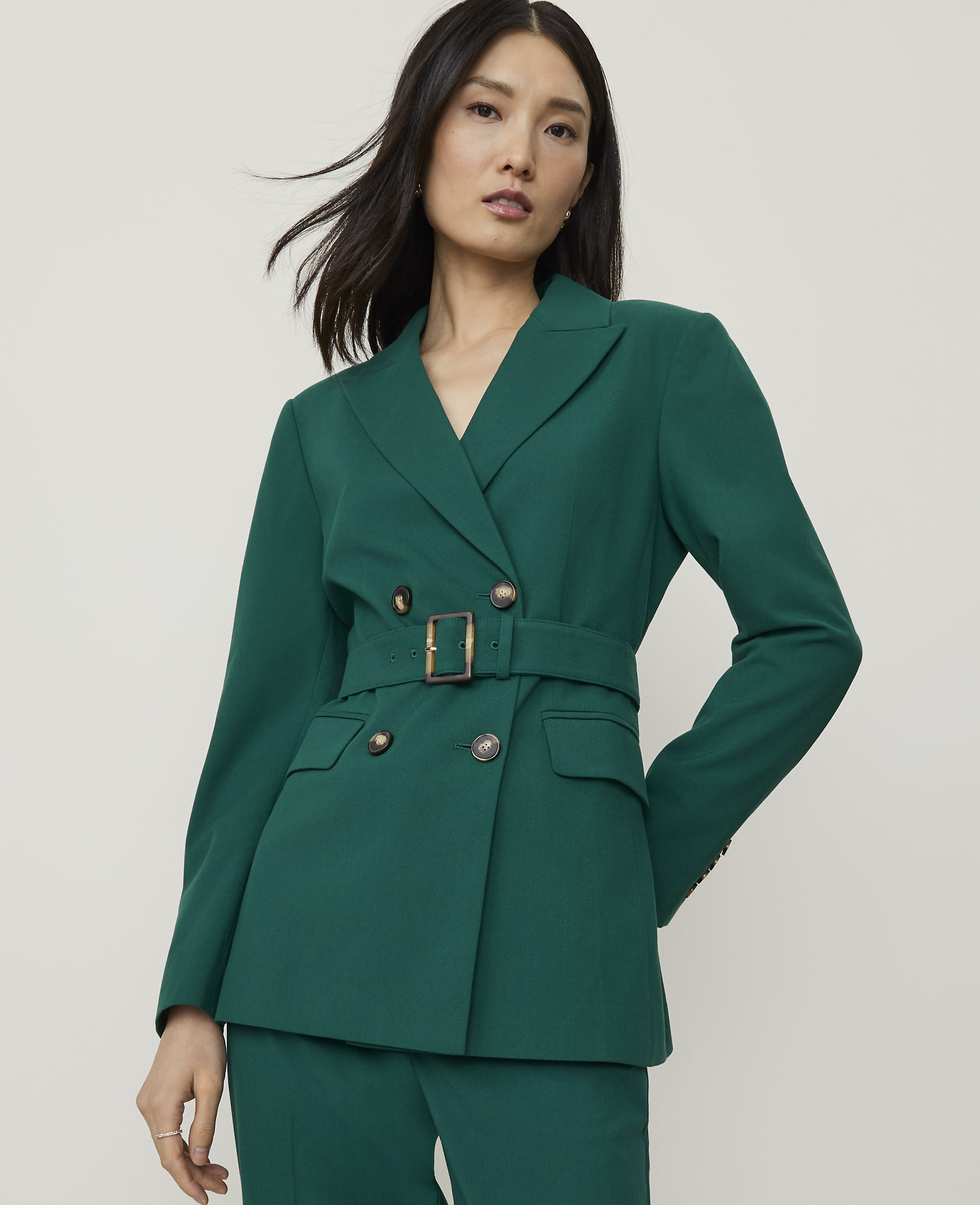 A woman in a green double-breasted blazer with a belt stands against a plain, light gray background.