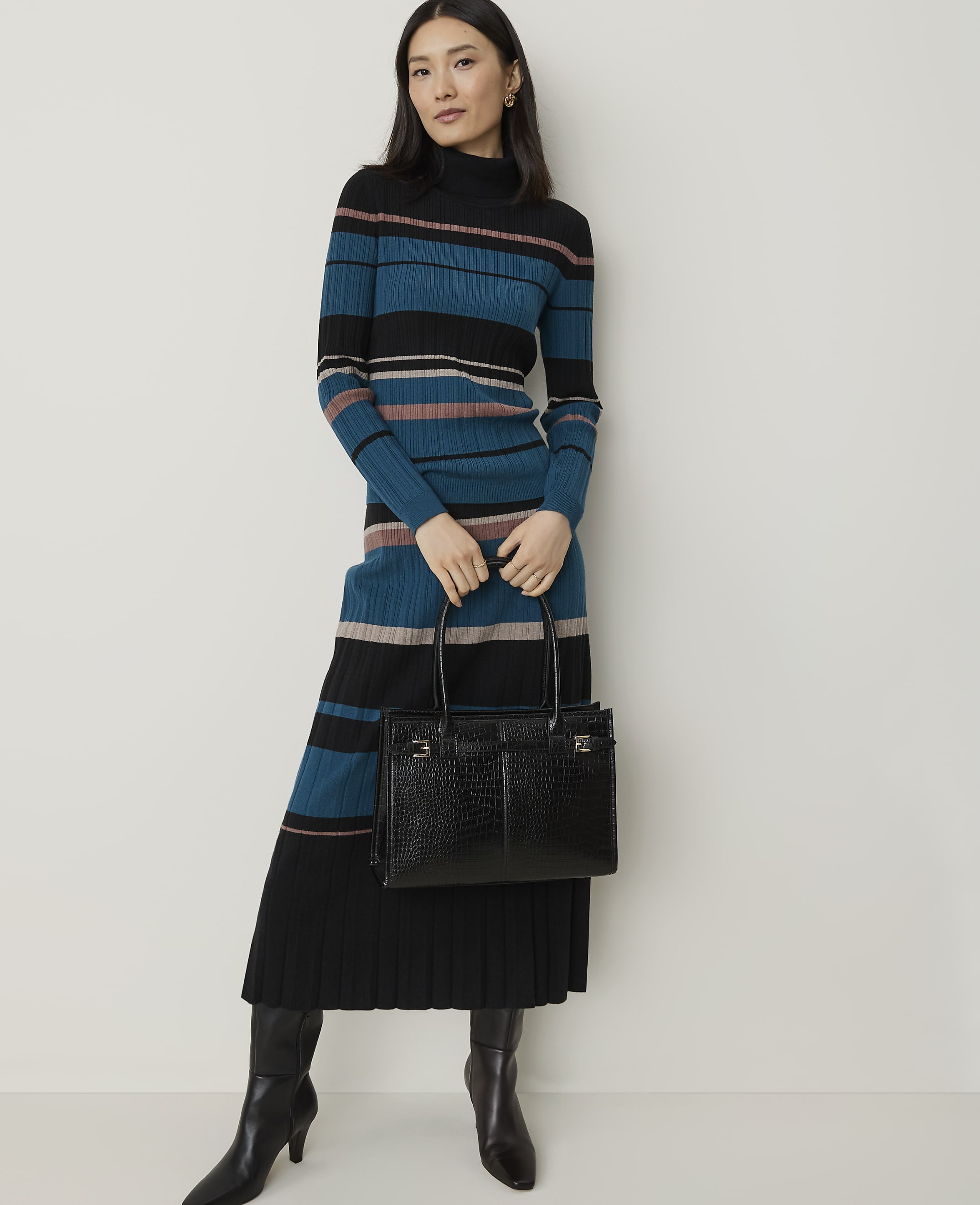 A woman in a striped turtleneck dress and black boots holds a black crocodile-embossed handbag against a plain white background.