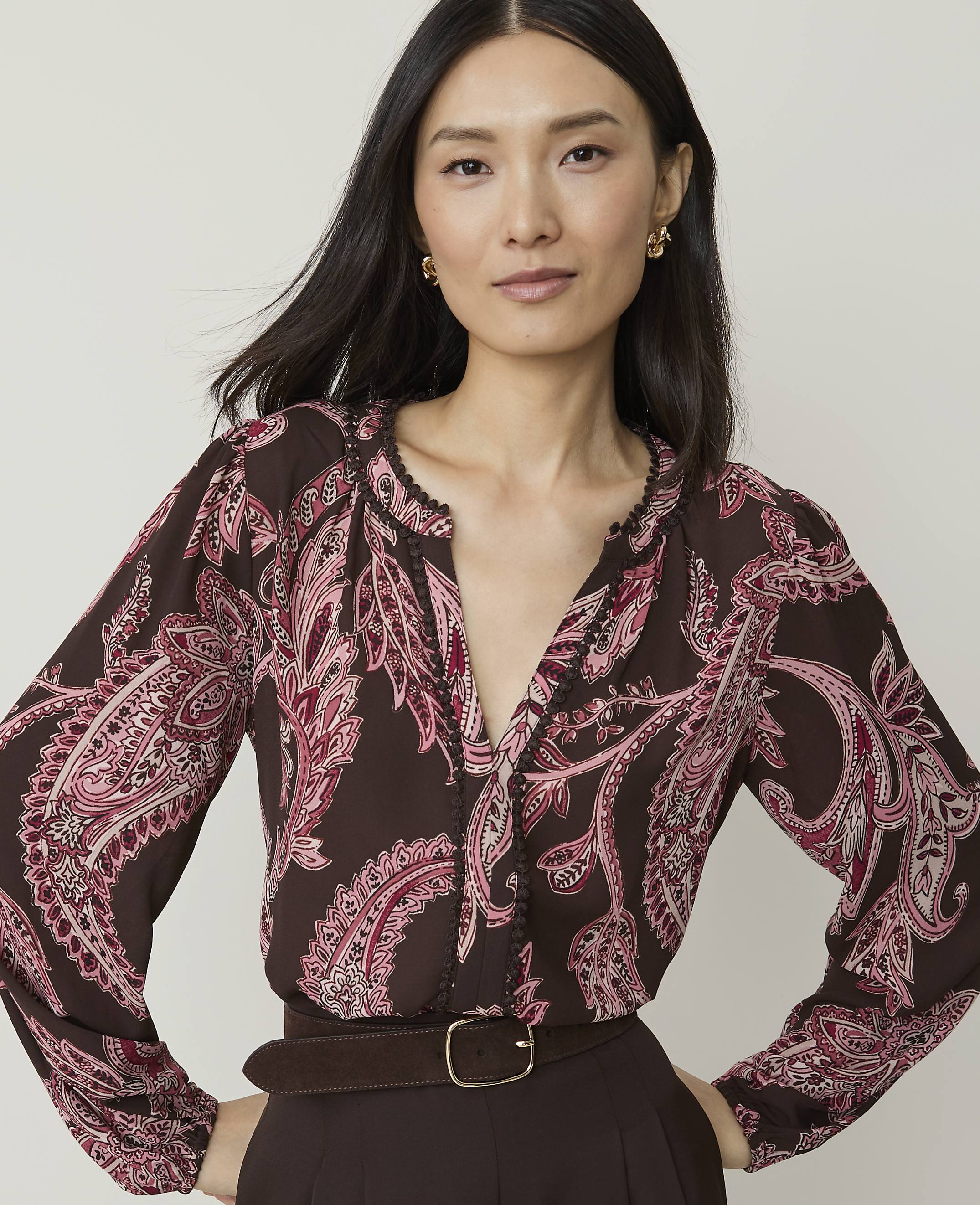 A woman with dark hair and a patterned blouse stands against a plain background.