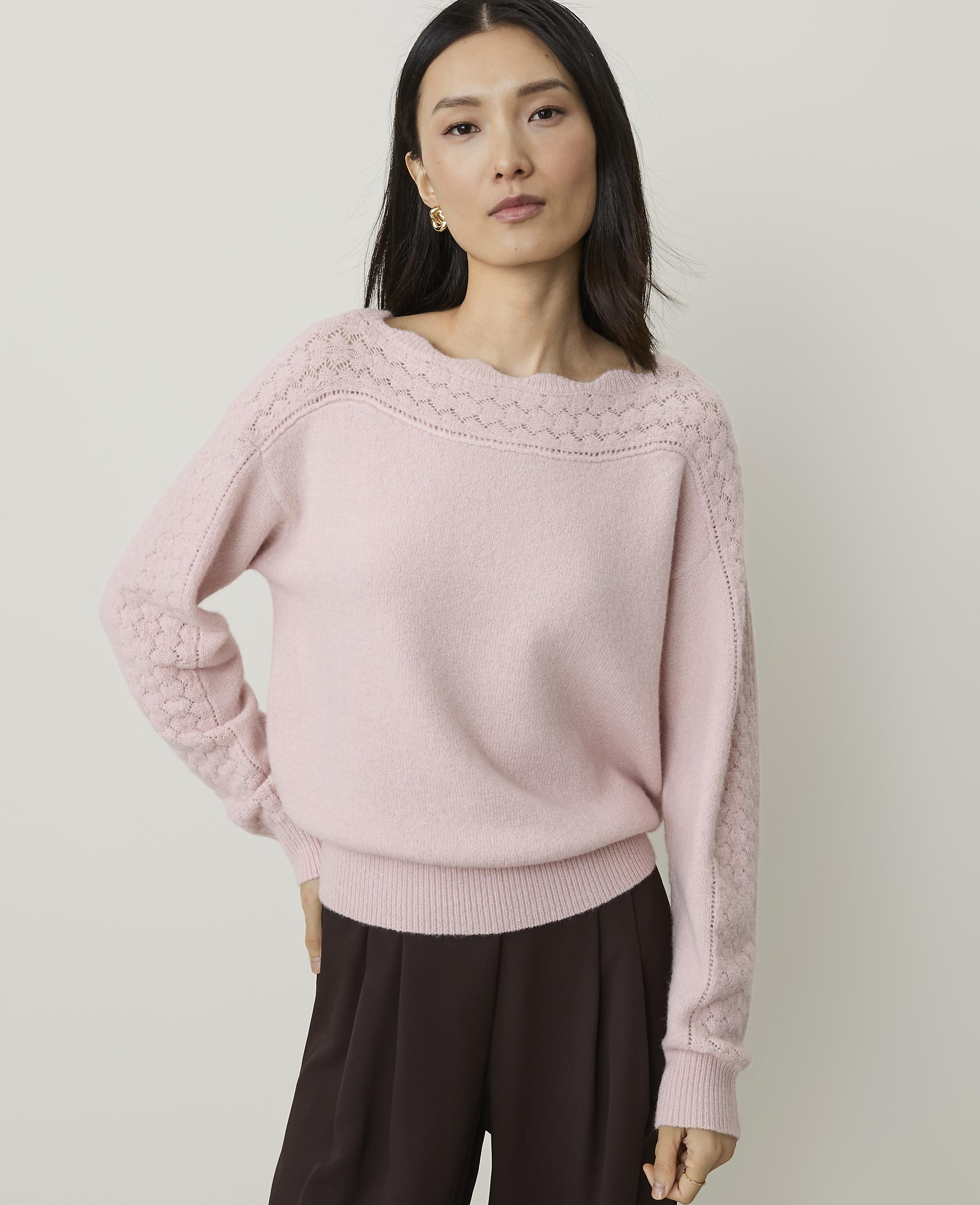 A woman in a pink sweater and brown pants stands against a plain background.