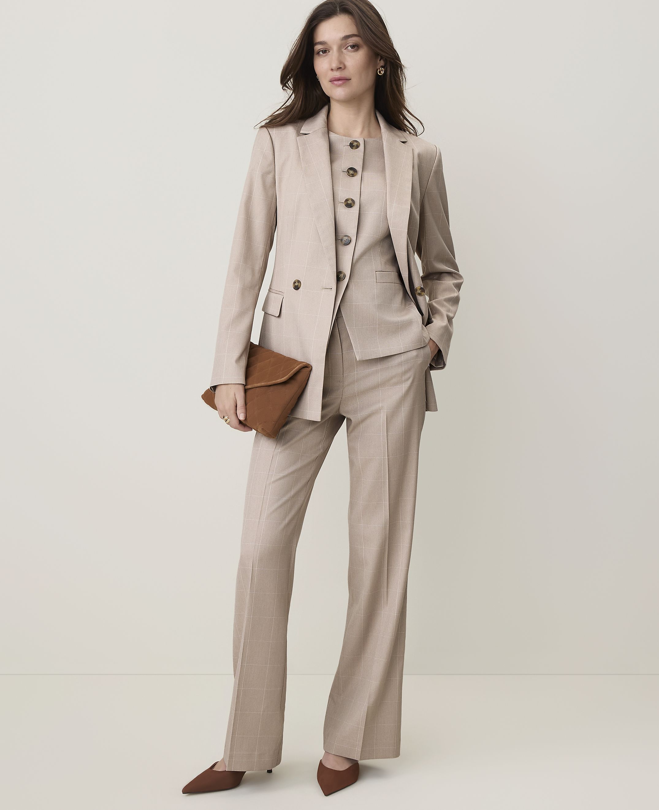A woman in a beige pantsuit and vest holds a brown clutch in front of a plain, light-colored background.
