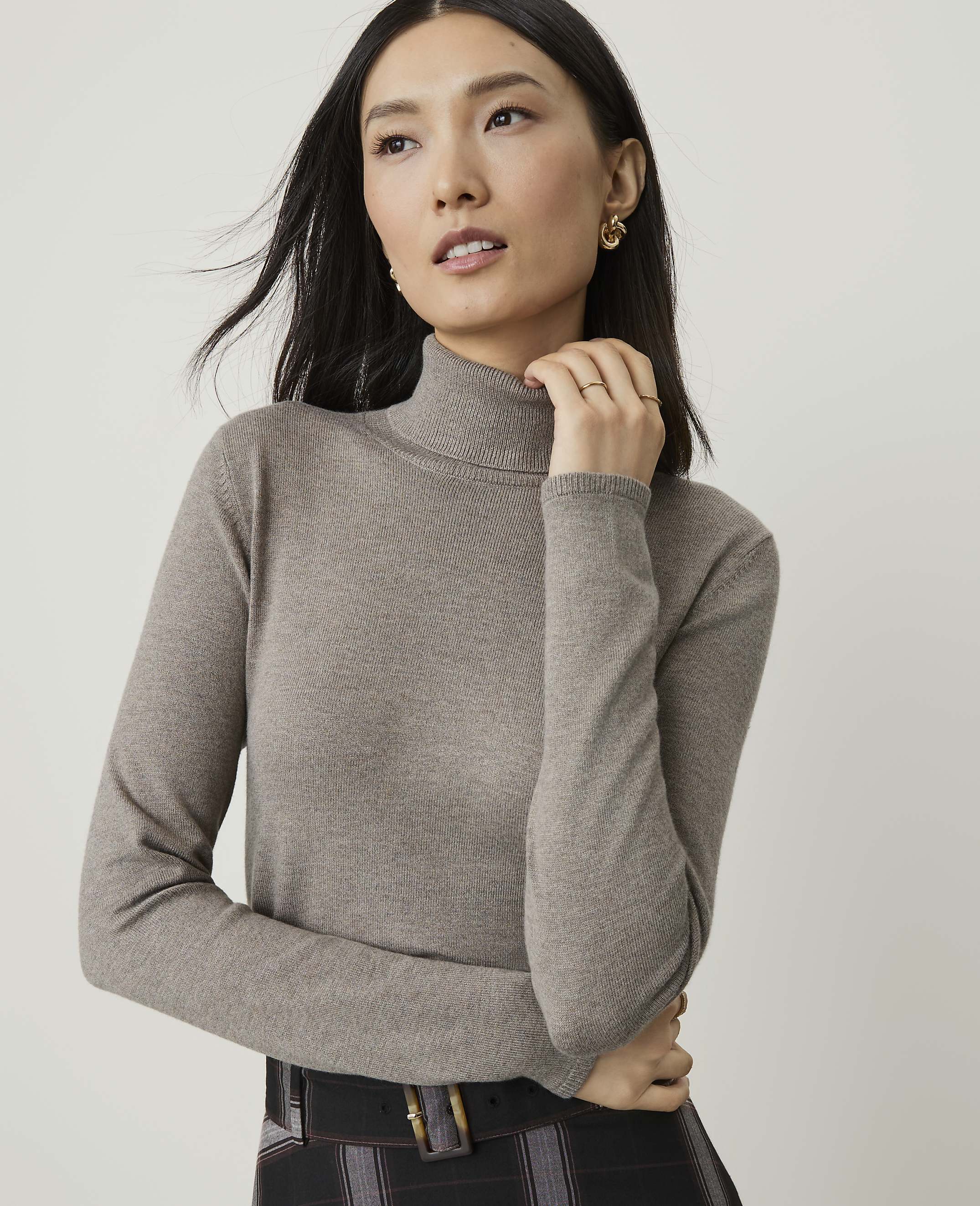 A woman in a taupe turtleneck sweater poses against a plain white background.