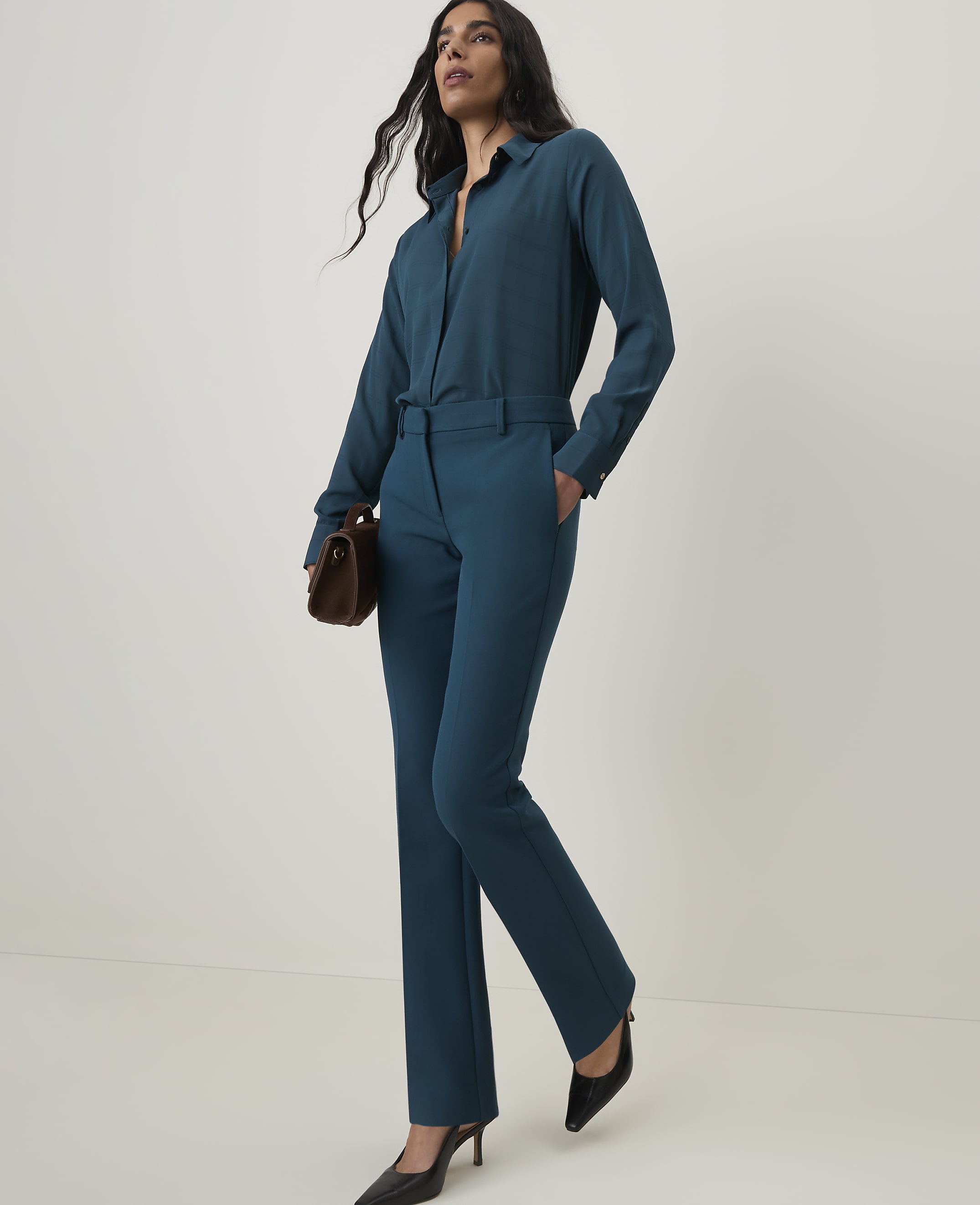 A woman in a teal pantsuit with a brown clutch stands against a plain white background.