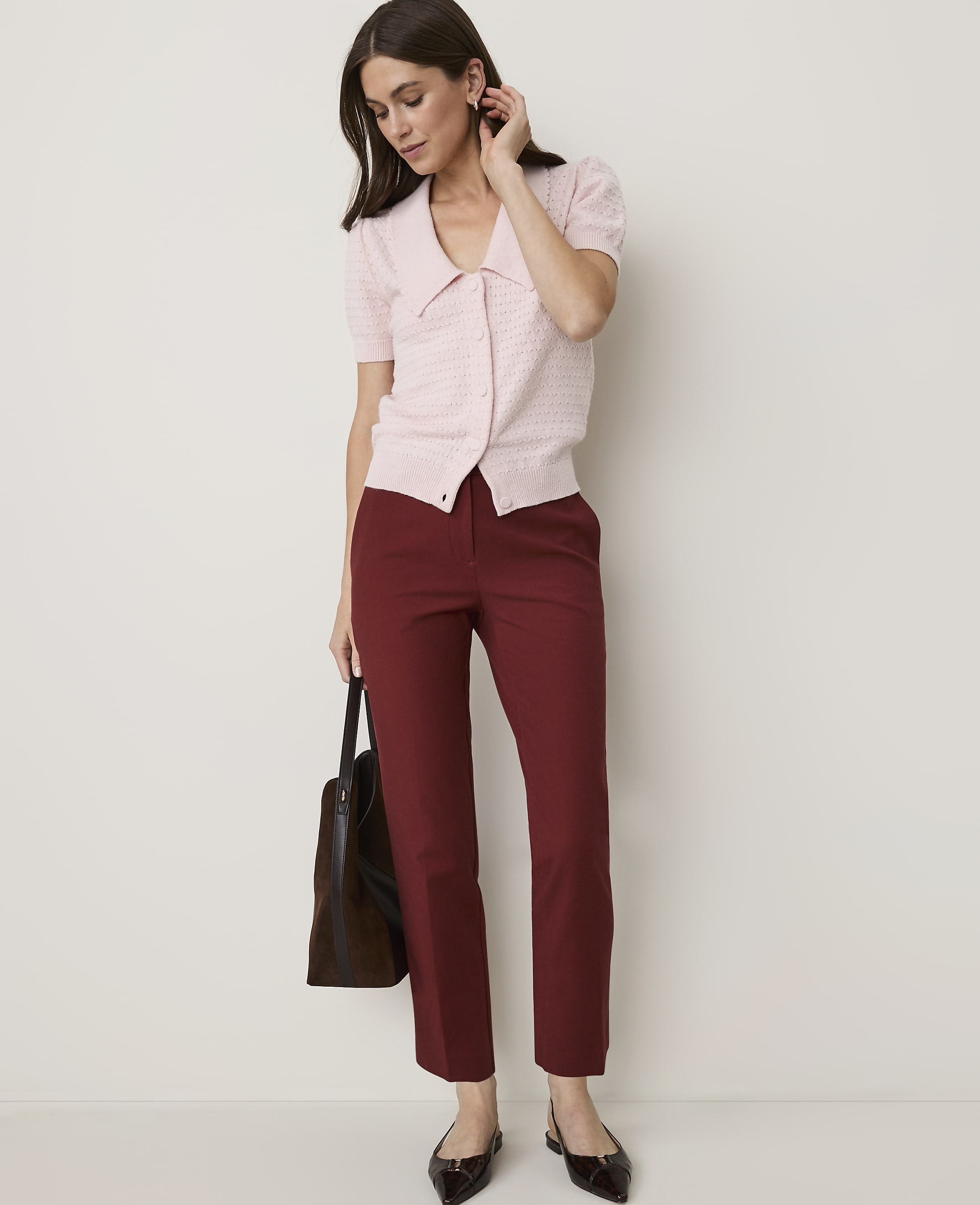 A woman in a pink cardigan and maroon pants stands against a plain white background, holding a dark tote bag.