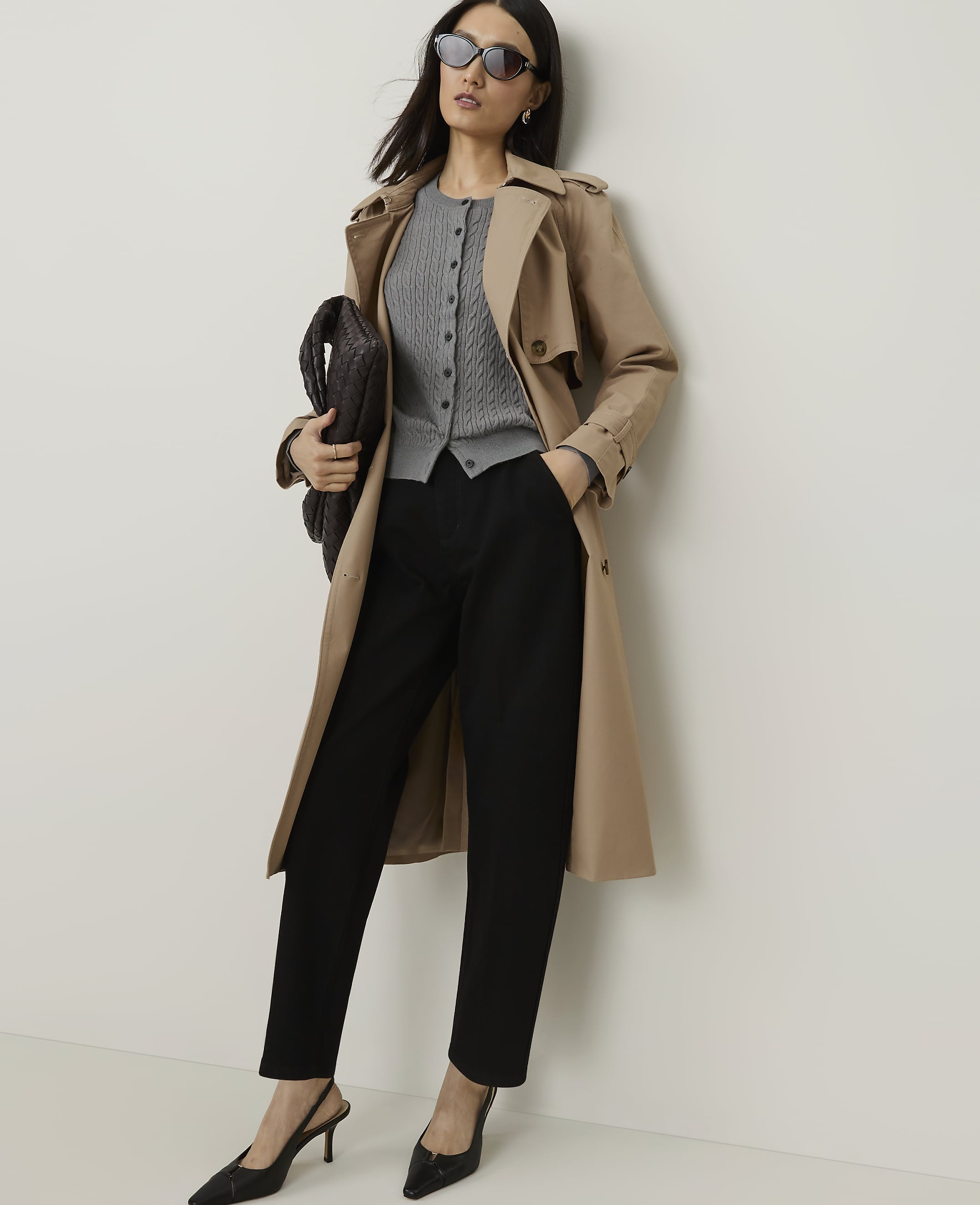 A woman in a trench coat, cardigan, and black pants poses against a plain white wall, holding a dark clutch.