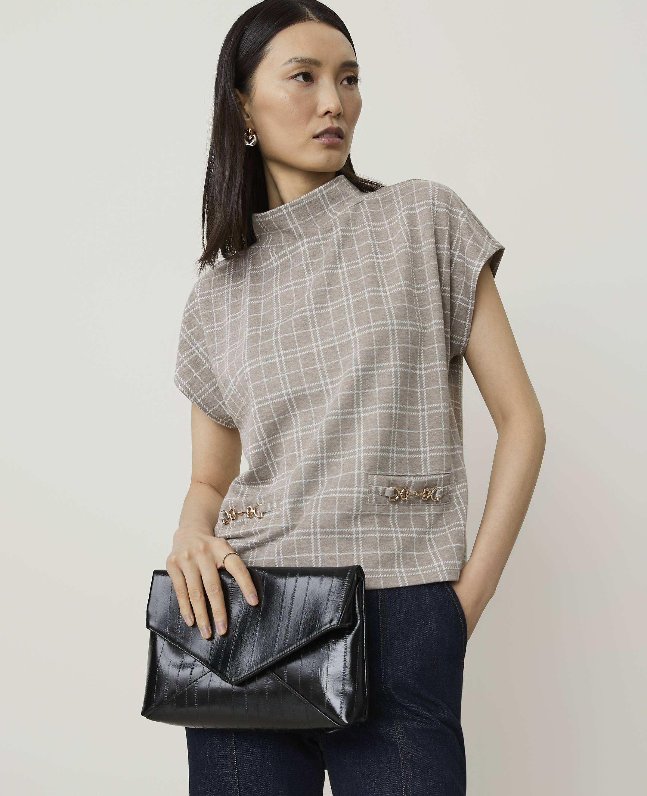 A woman in a plaid top and dark pants holds a black clutch in front of a plain background.