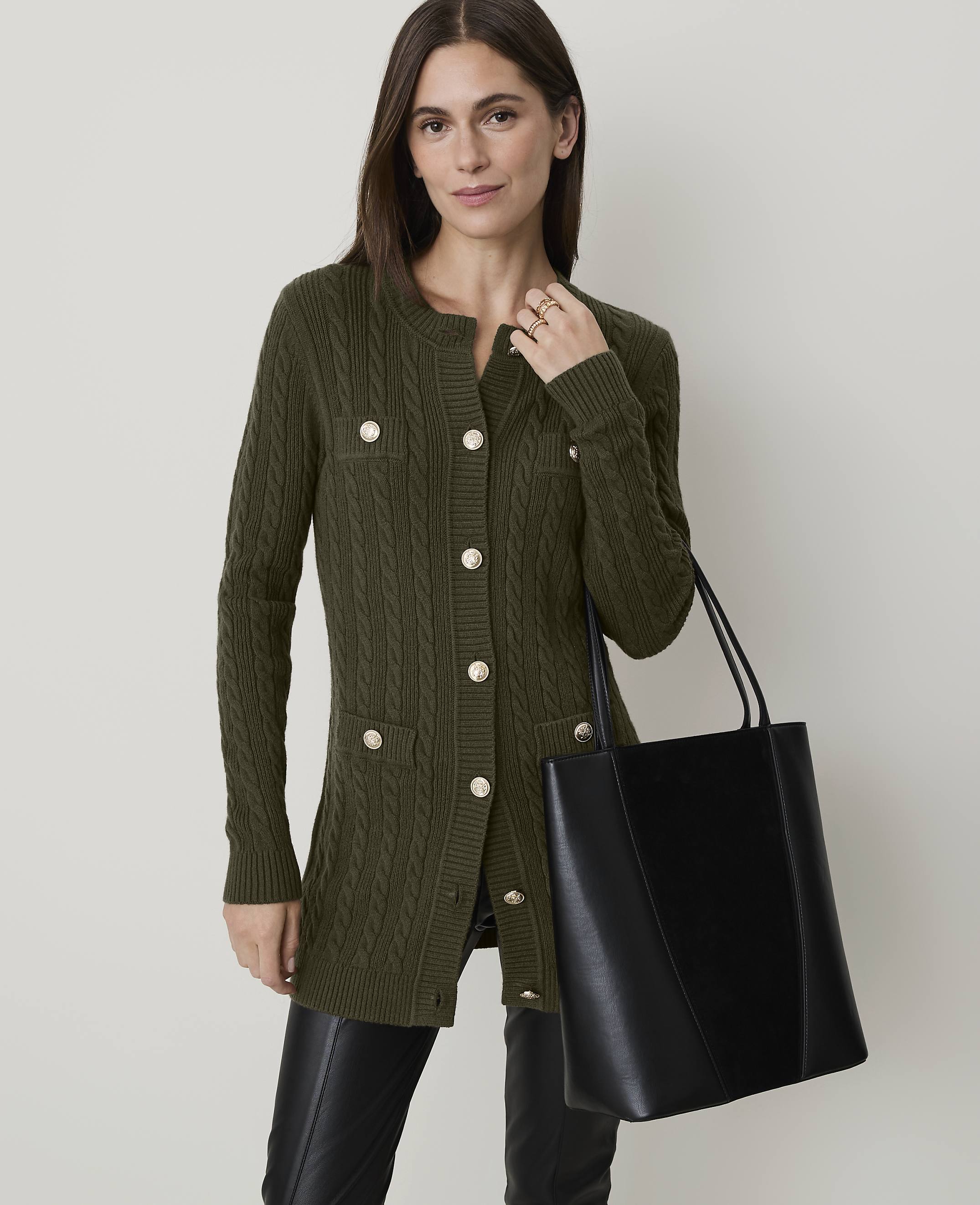 A woman in a brown cable-knit cardigan and black pants stands with a black tote bag in front of a plain, light-colored background.