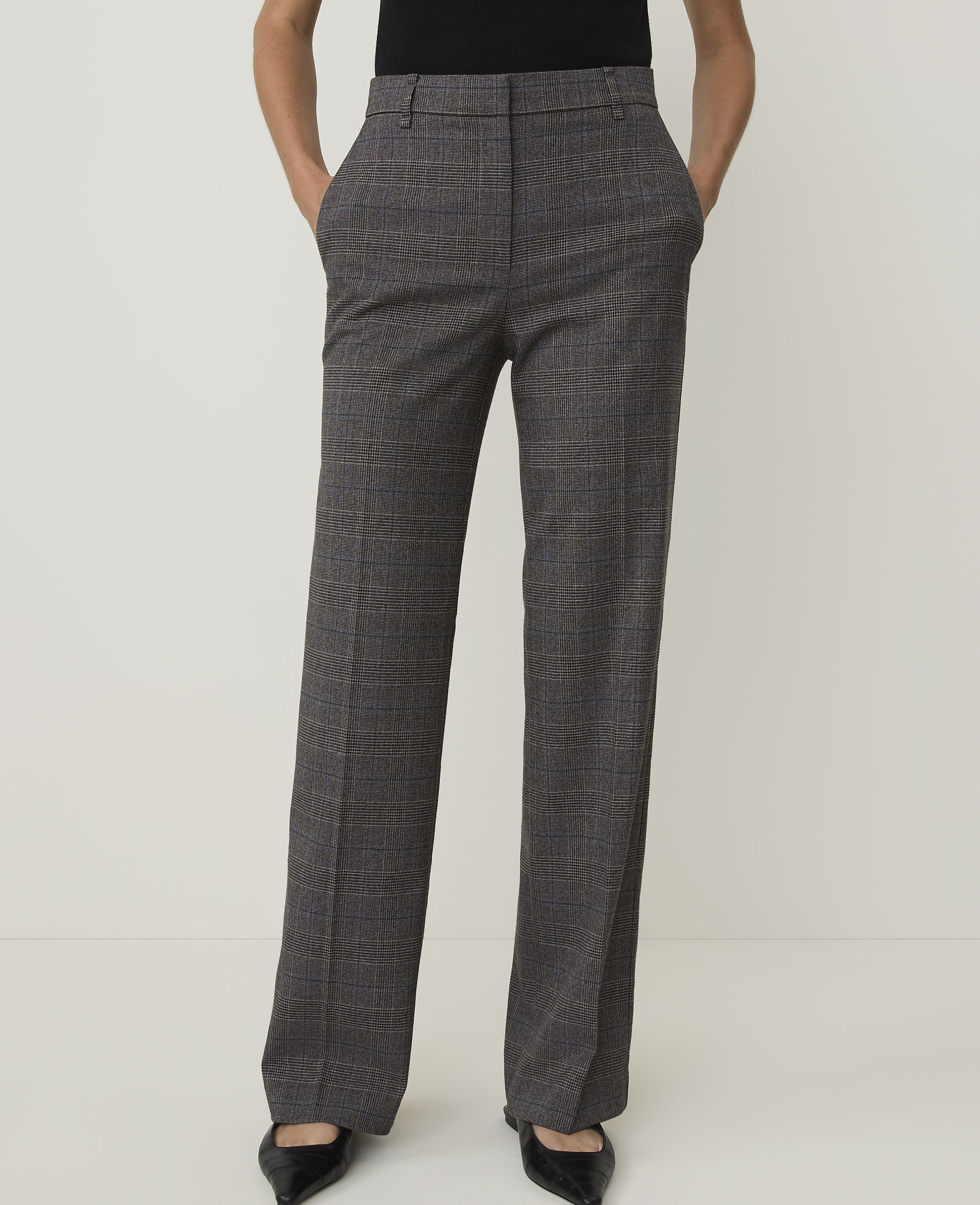 A person stands in front of a plain white background, wearing plaid trousers and black pointed-toe shoes.