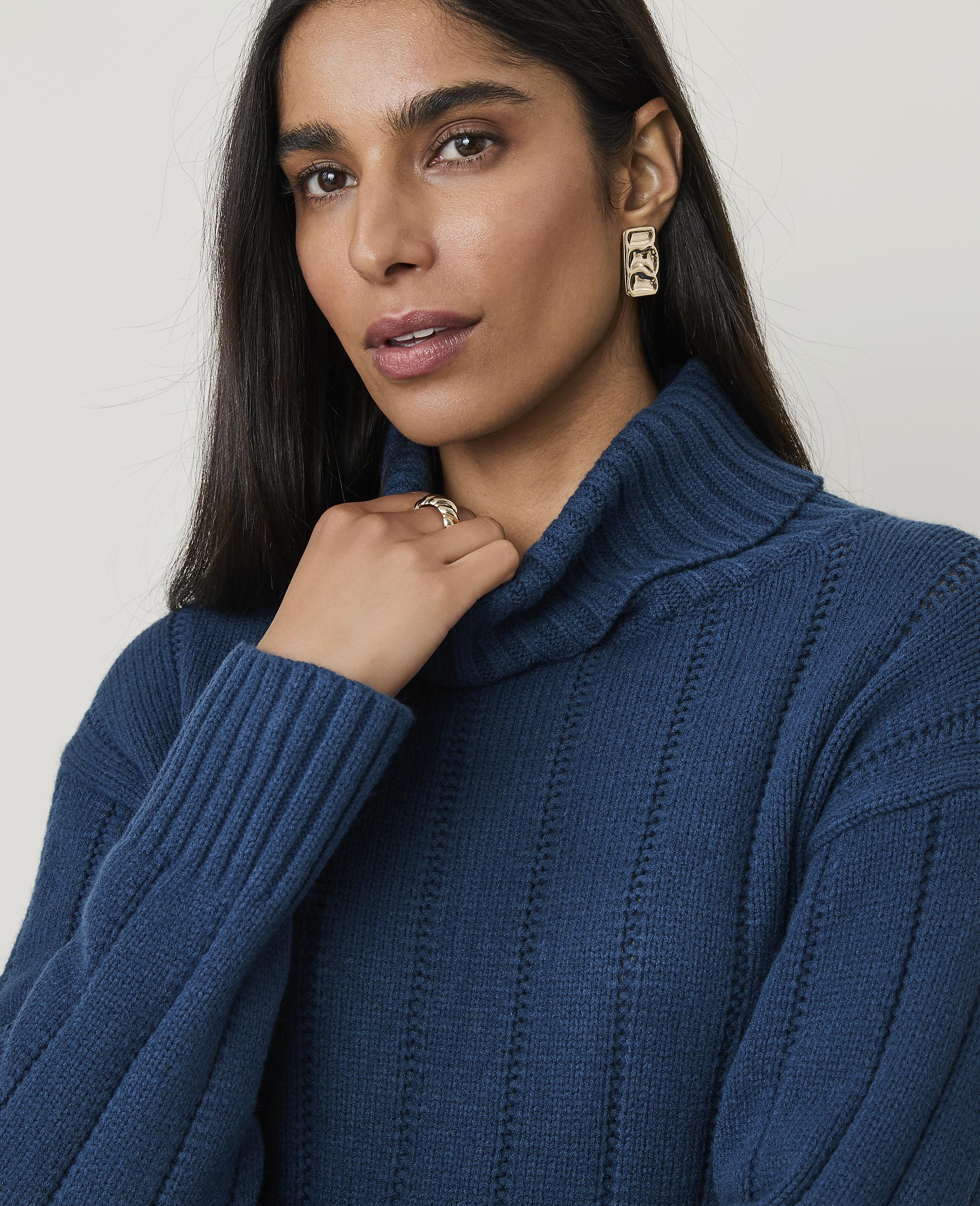 A woman with dark hair and a teal turtleneck sweater is in the foreground, while a plain, light-colored background is visible behind her.