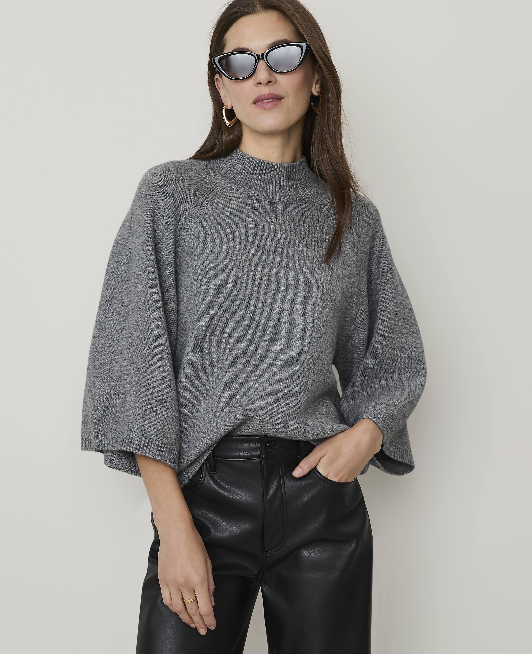 A woman in a grey sweater and black leather pants poses against a plain background.