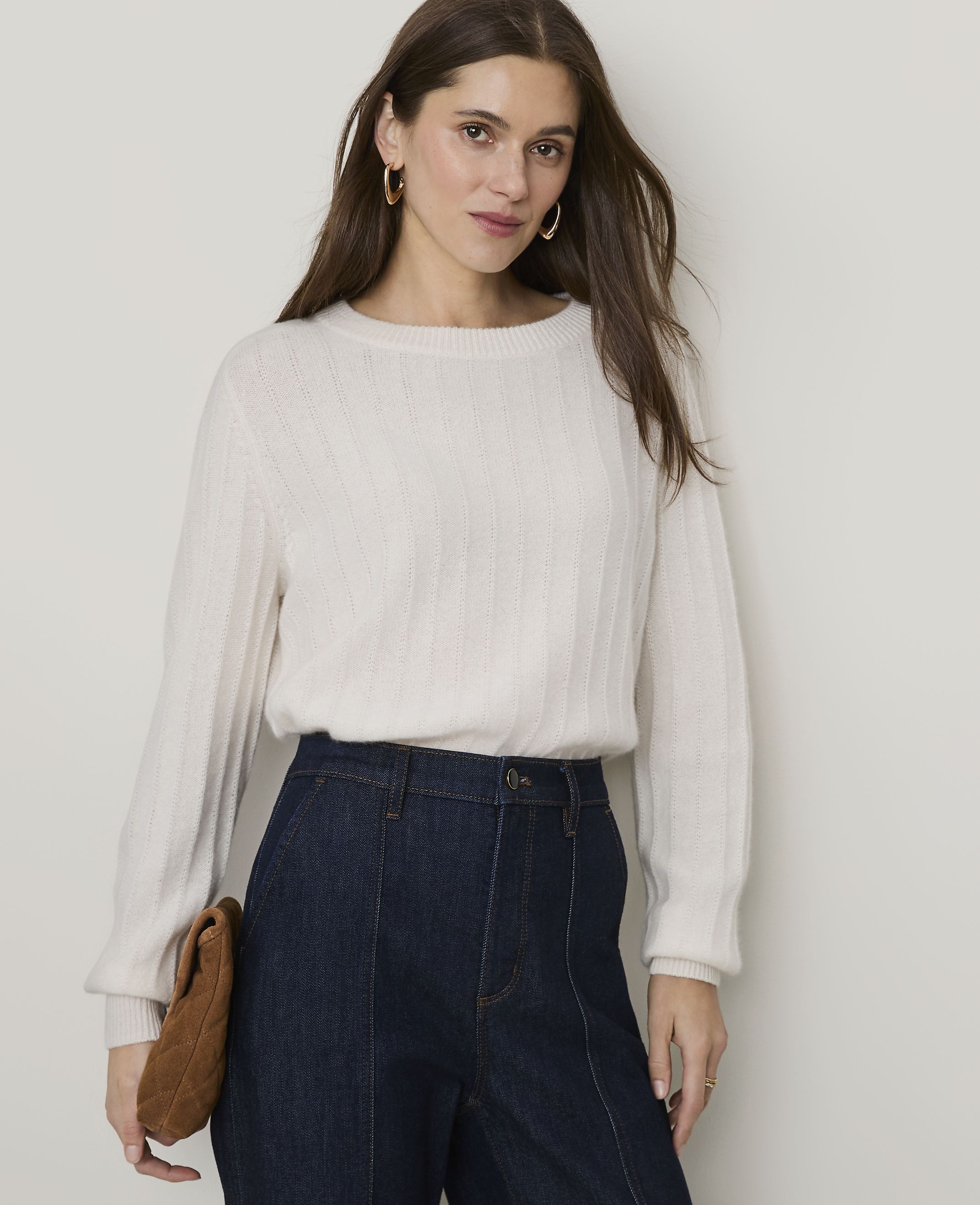 A woman in a cream sweater and dark jeans stands against a plain, light-colored background, holding a brown clutch.
