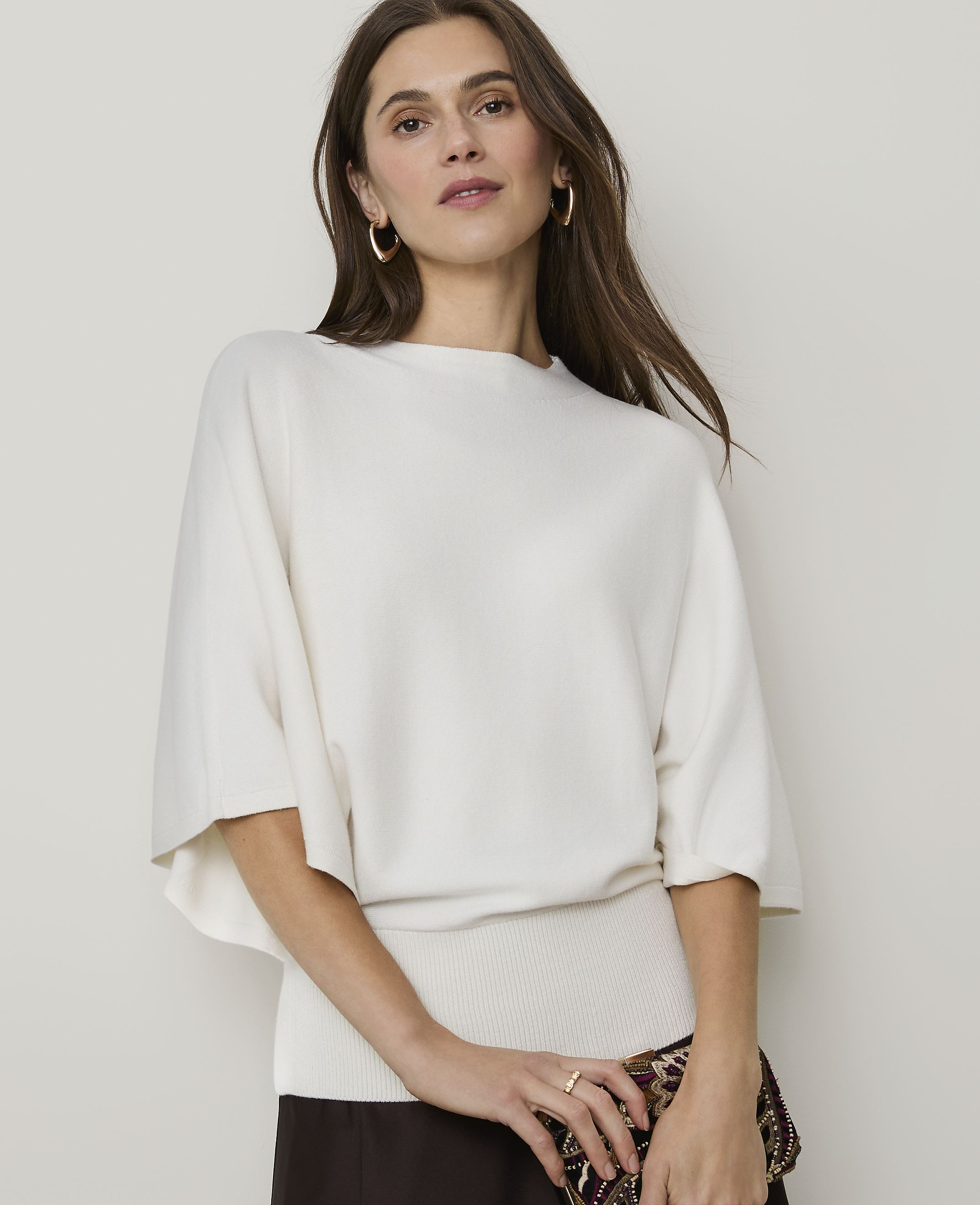 A woman in a cream-colored sweater and dark skirt poses against a plain, light background.