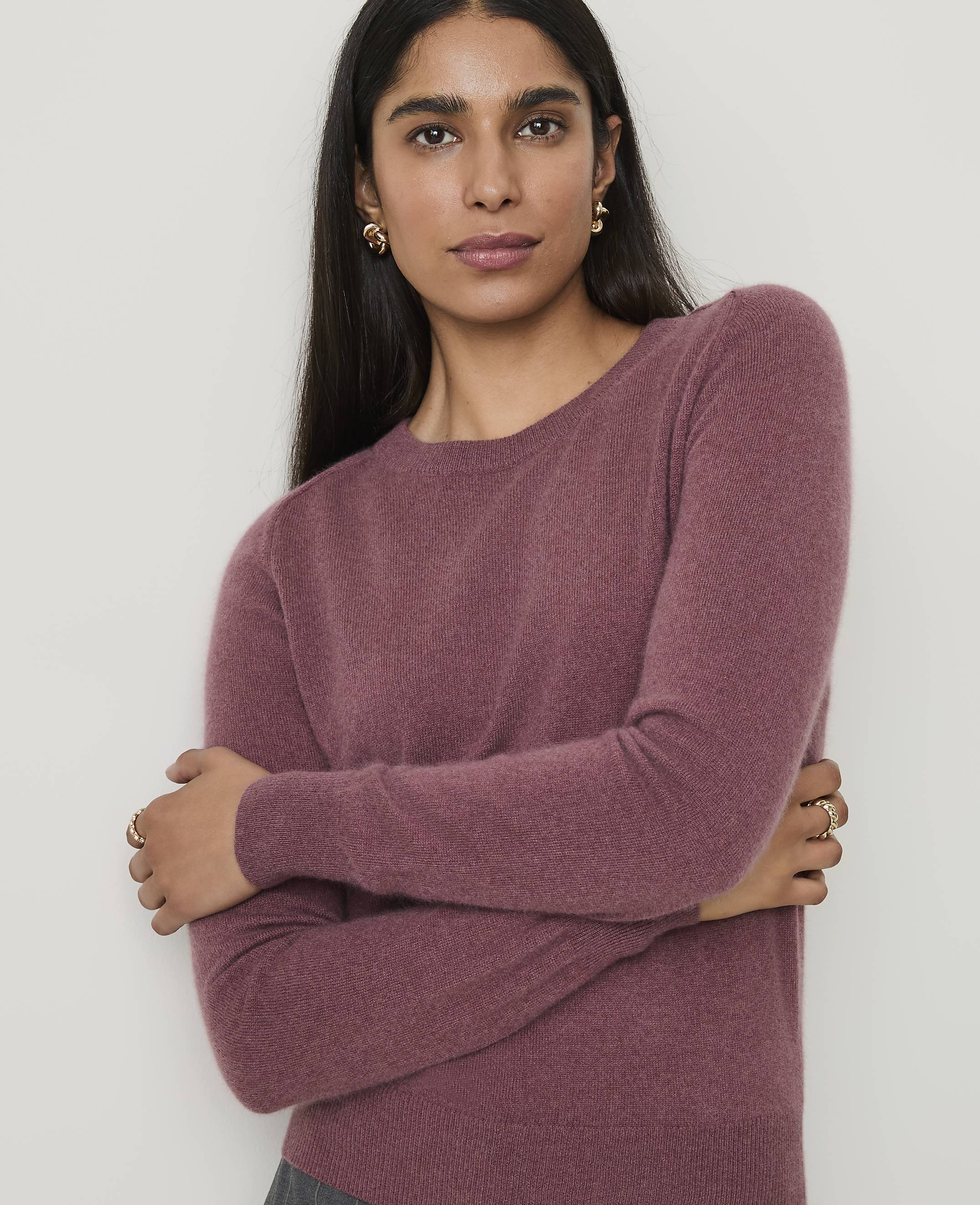 A woman in a mauve sweater crosses her arms in front of a plain white background.