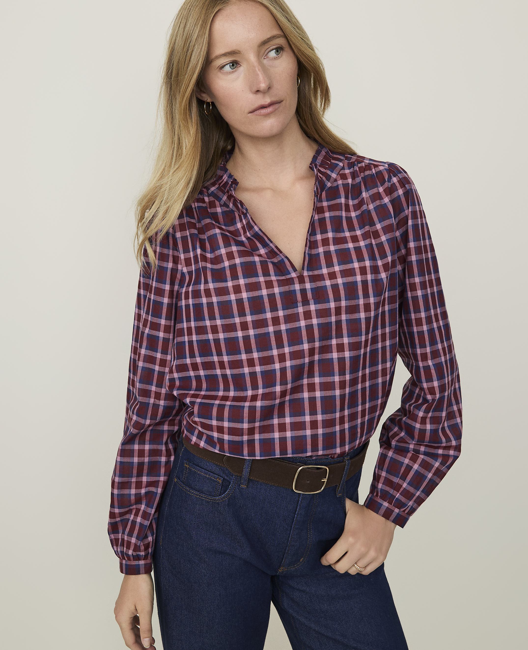 A woman in a maroon and blue plaid blouse and dark jeans stands against a plain, light background.