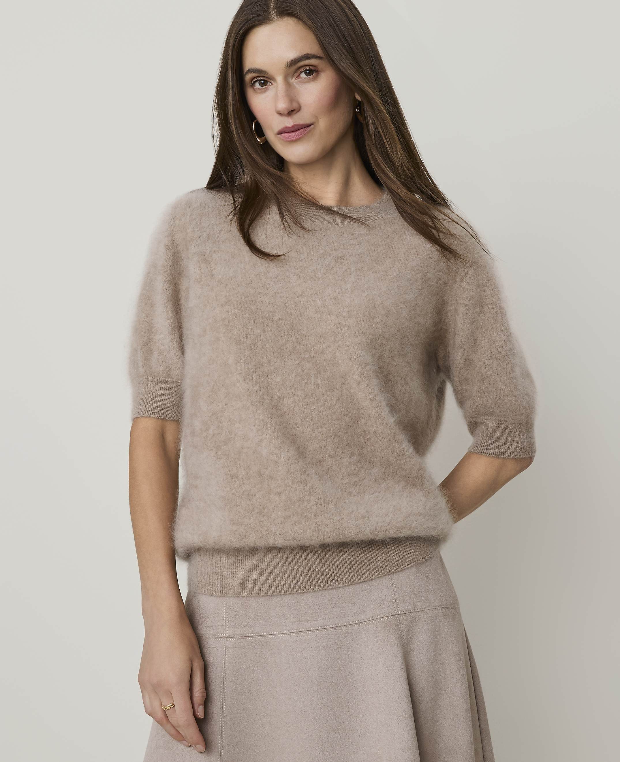 A woman in a fuzzy beige sweater and skirt stands against a plain, light background.
