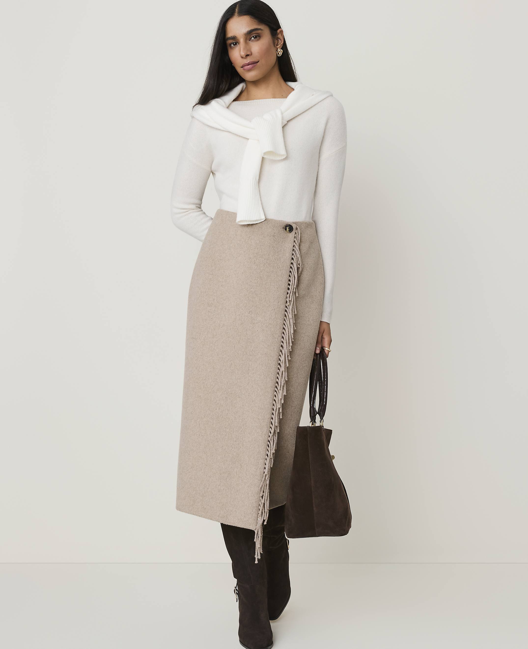 A woman in a cream sweater and beige skirt stands against a plain, light background, holding a dark brown bag.