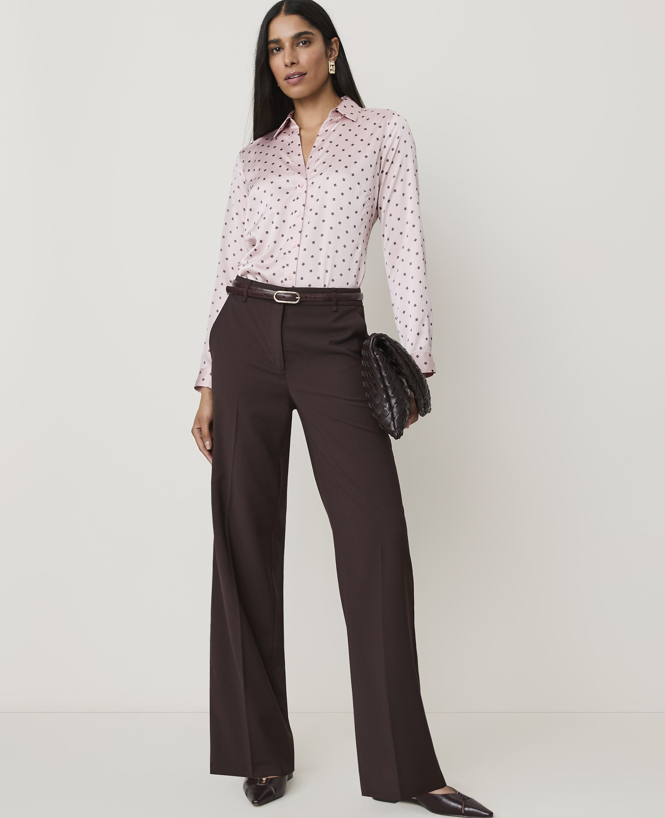 A woman in a pink polka dot blouse and brown wide-leg pants stands against a plain white background, holding a woven clutch.