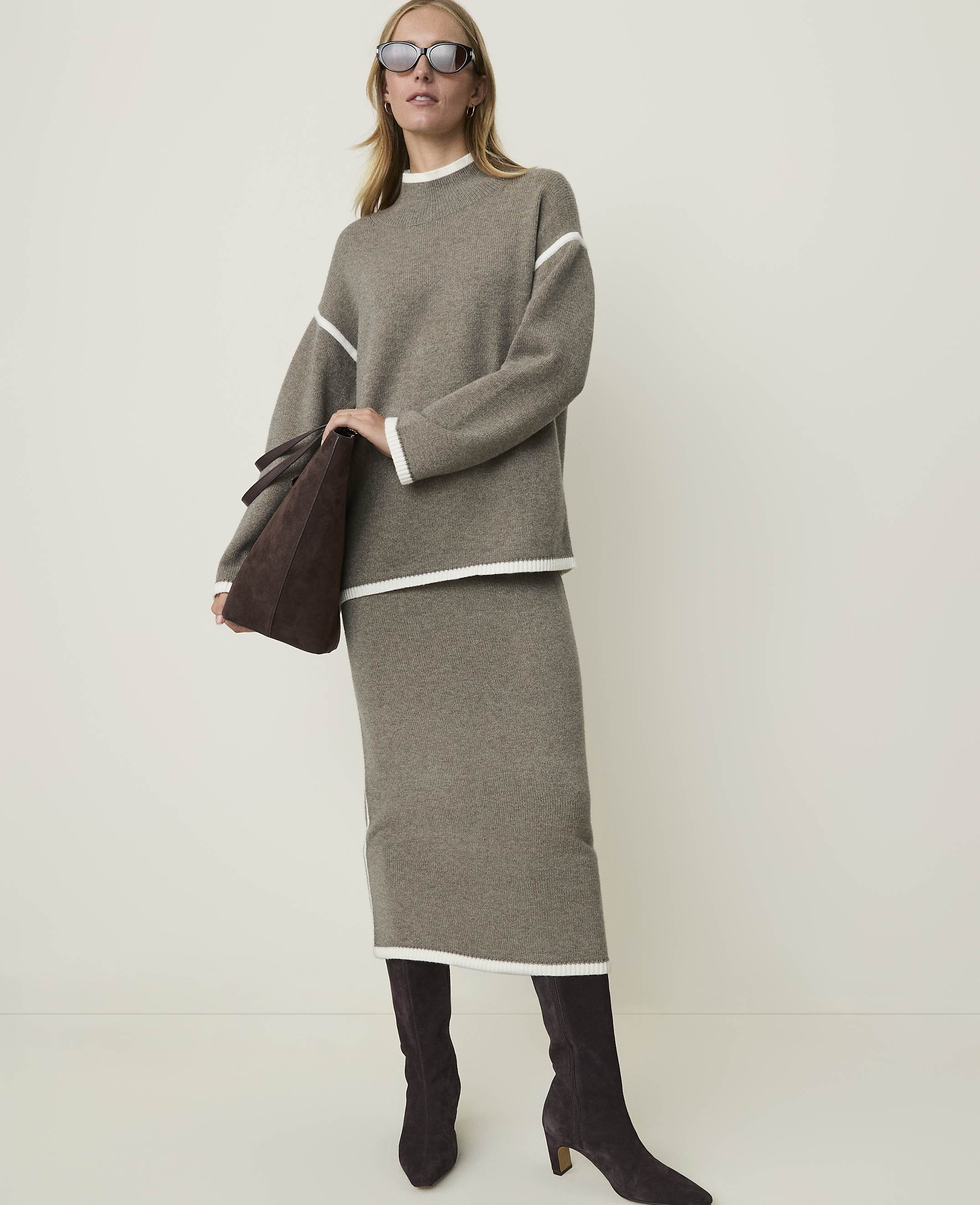 A woman in a taupe knit sweater and skirt with white trim, holding a brown suede tote bag, stands against a plain, light-colored background.