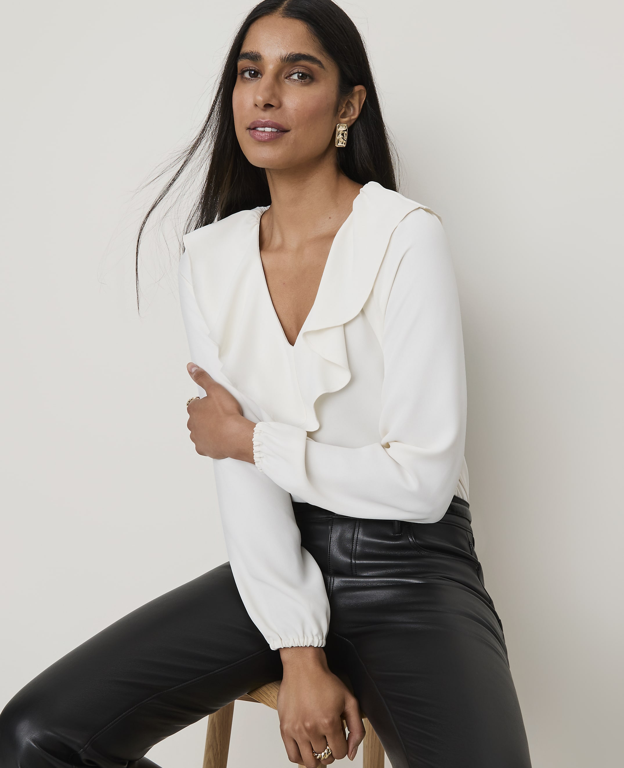 A woman in a cream ruffled blouse and black leather pants poses against a plain, light-colored background.