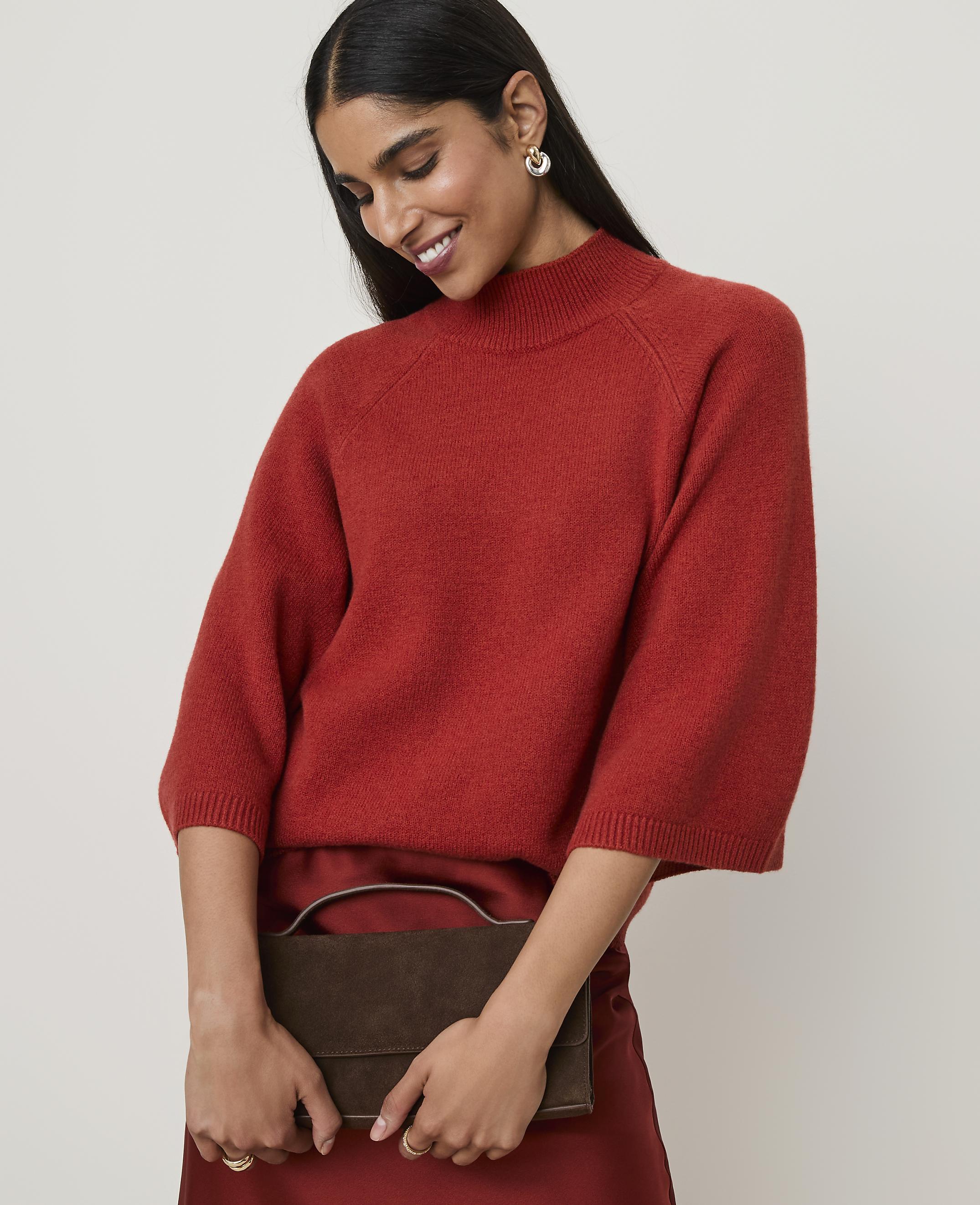 A woman in a rust-colored sweater and skirt holds a dark brown clutch against a plain beige background.