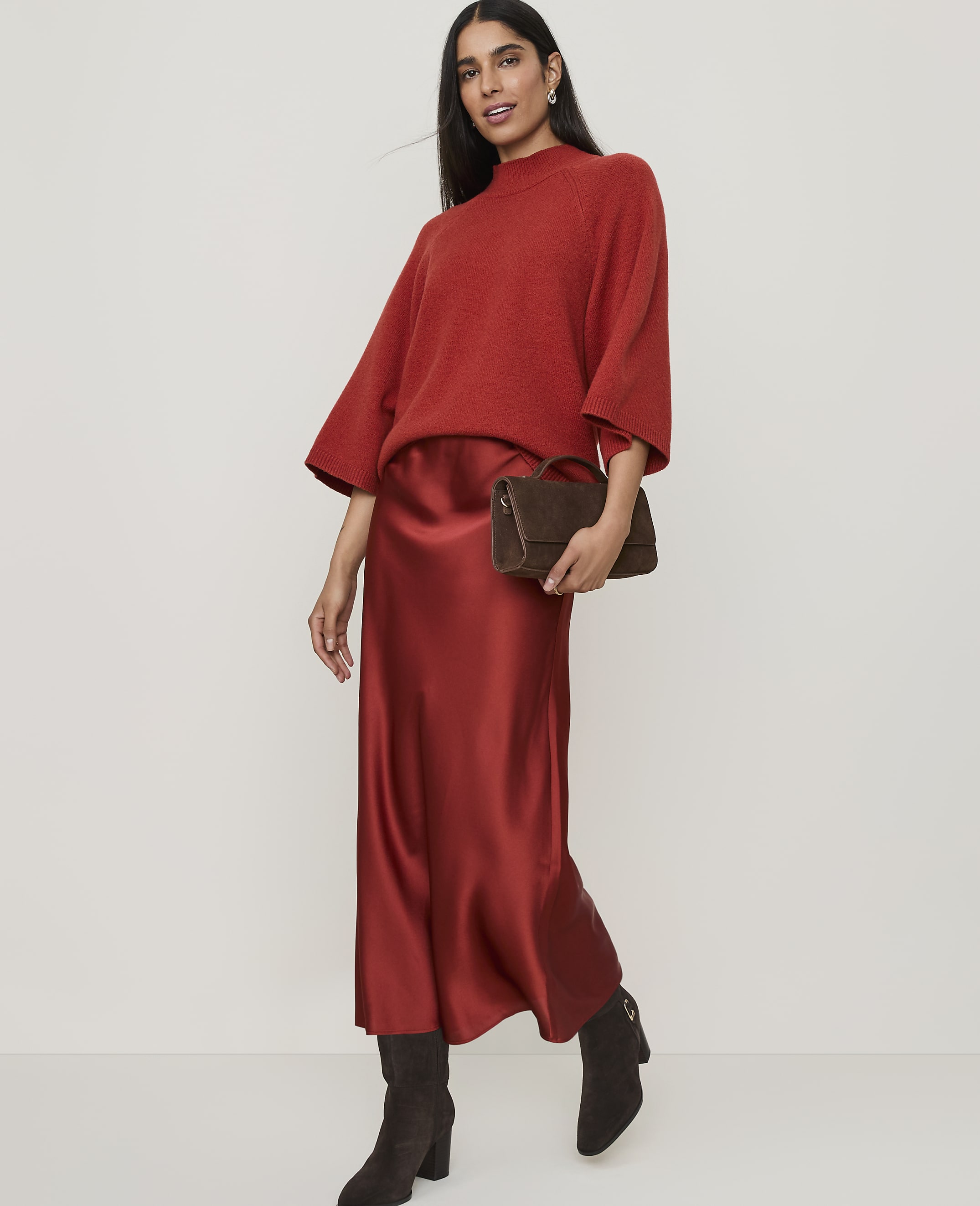 A woman in a rust-colored sweater and satin skirt poses against a plain, light background.
