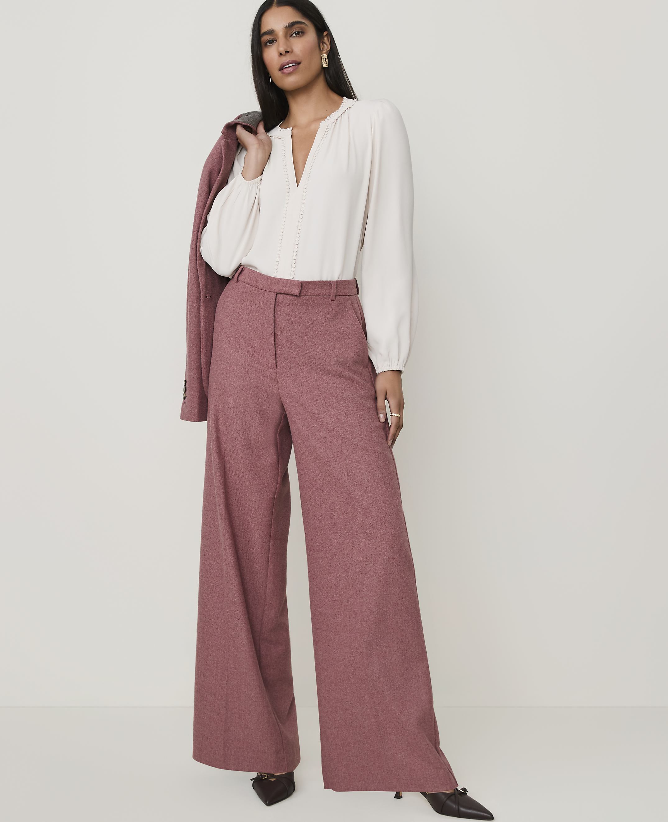 A woman in a cream blouse and wide-leg pink pants stands against a plain white background, with a pink blazer draped over her shoulder.