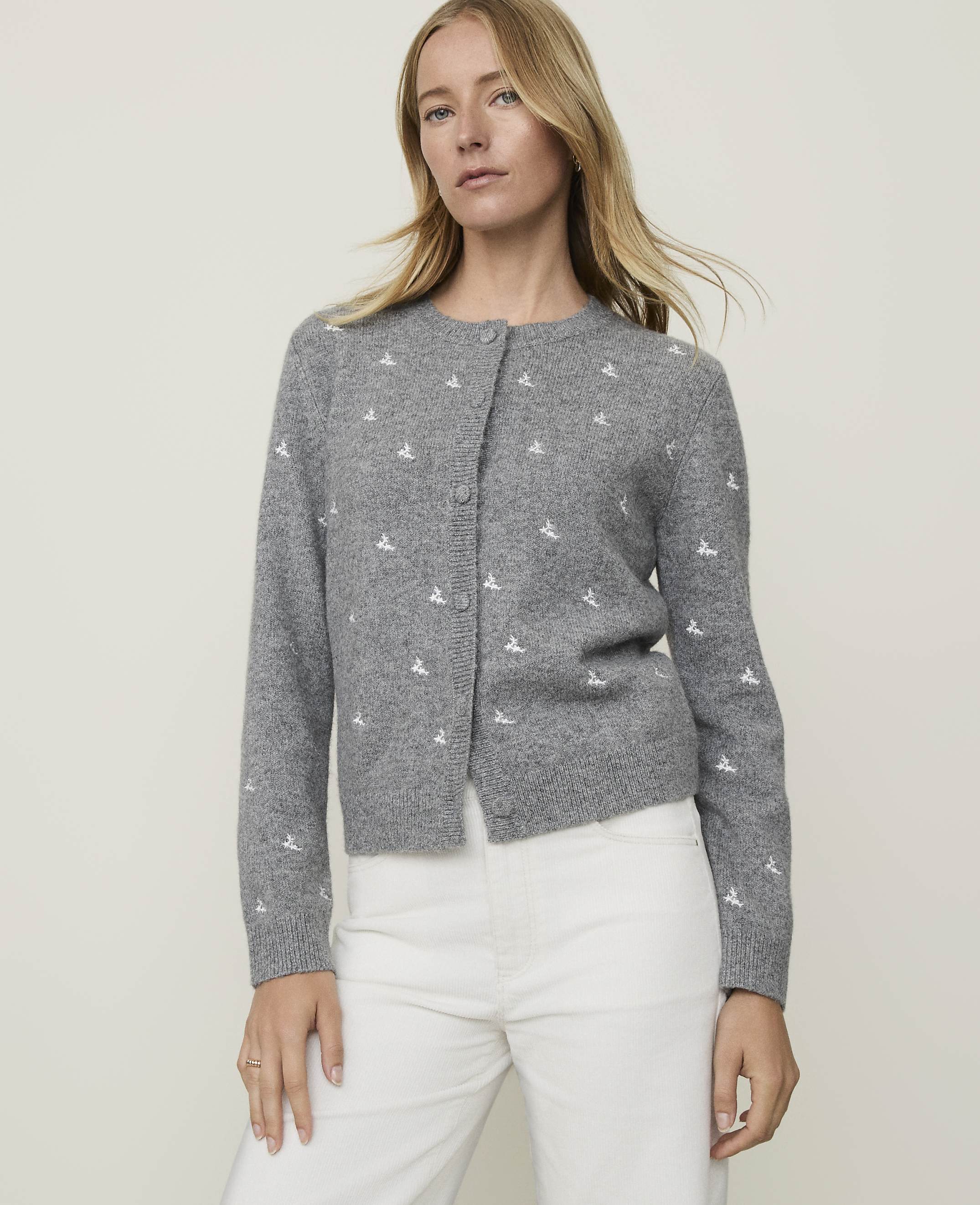 A woman in a grey cardigan with a white embroidered pattern stands against a plain grey background.