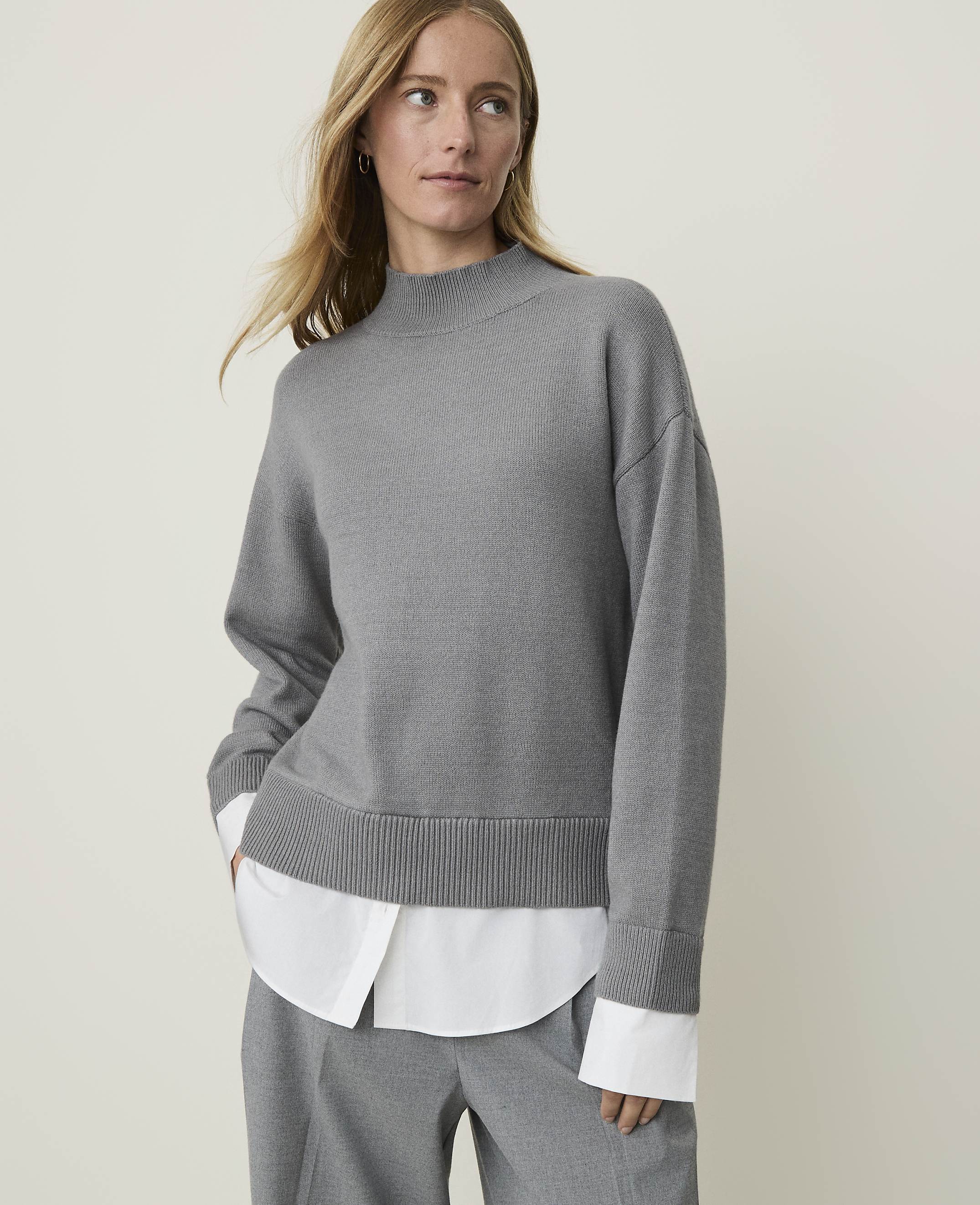 A woman in a grey mock-neck sweater layered over a white collared shirt stands against a plain, light grey background.