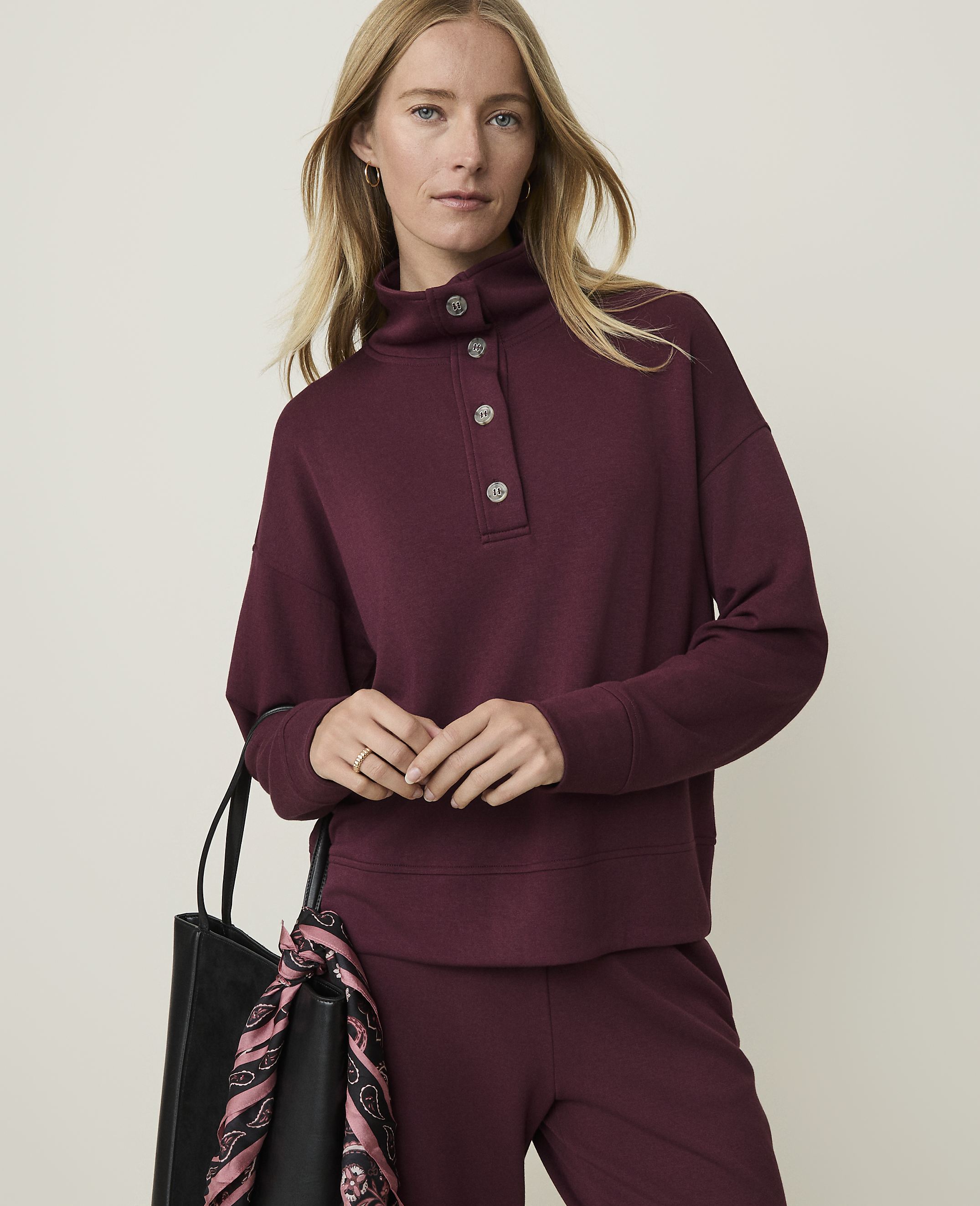A woman in a burgundy mock-neck sweatshirt and matching pants stands against a plain, light background, with a black tote bag at her side.