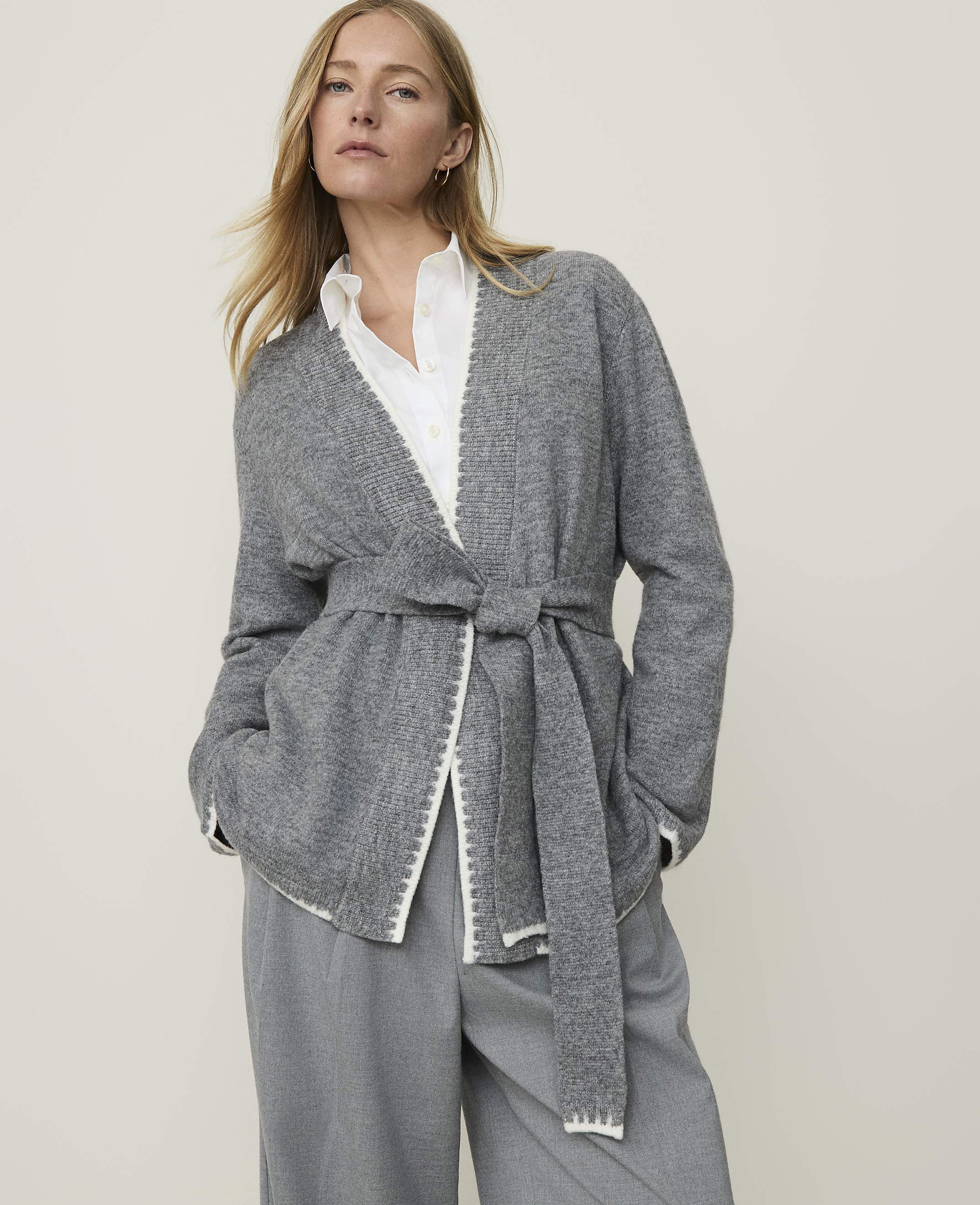 A woman in a grey cardigan and grey pants stands against a plain, light grey background.
