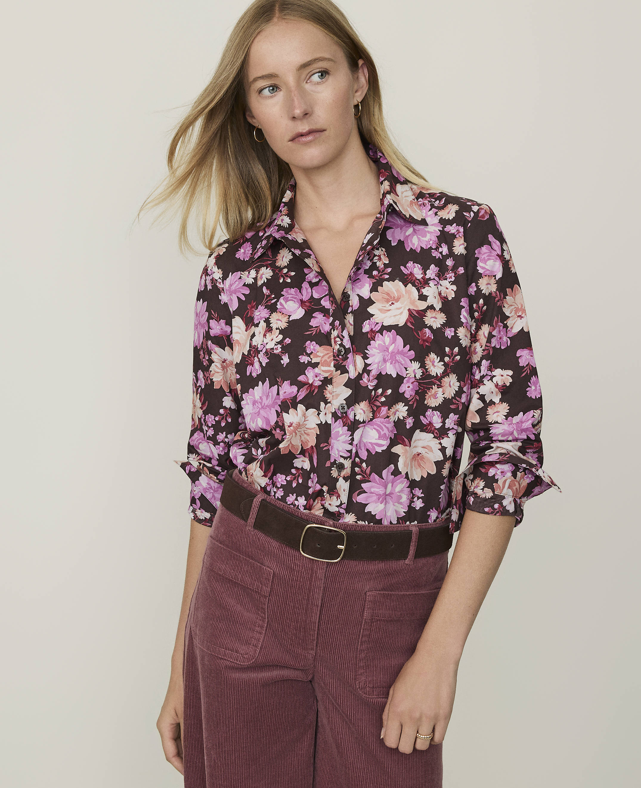 A woman in a floral shirt and maroon corduroy pants stands against a plain, light-colored background.