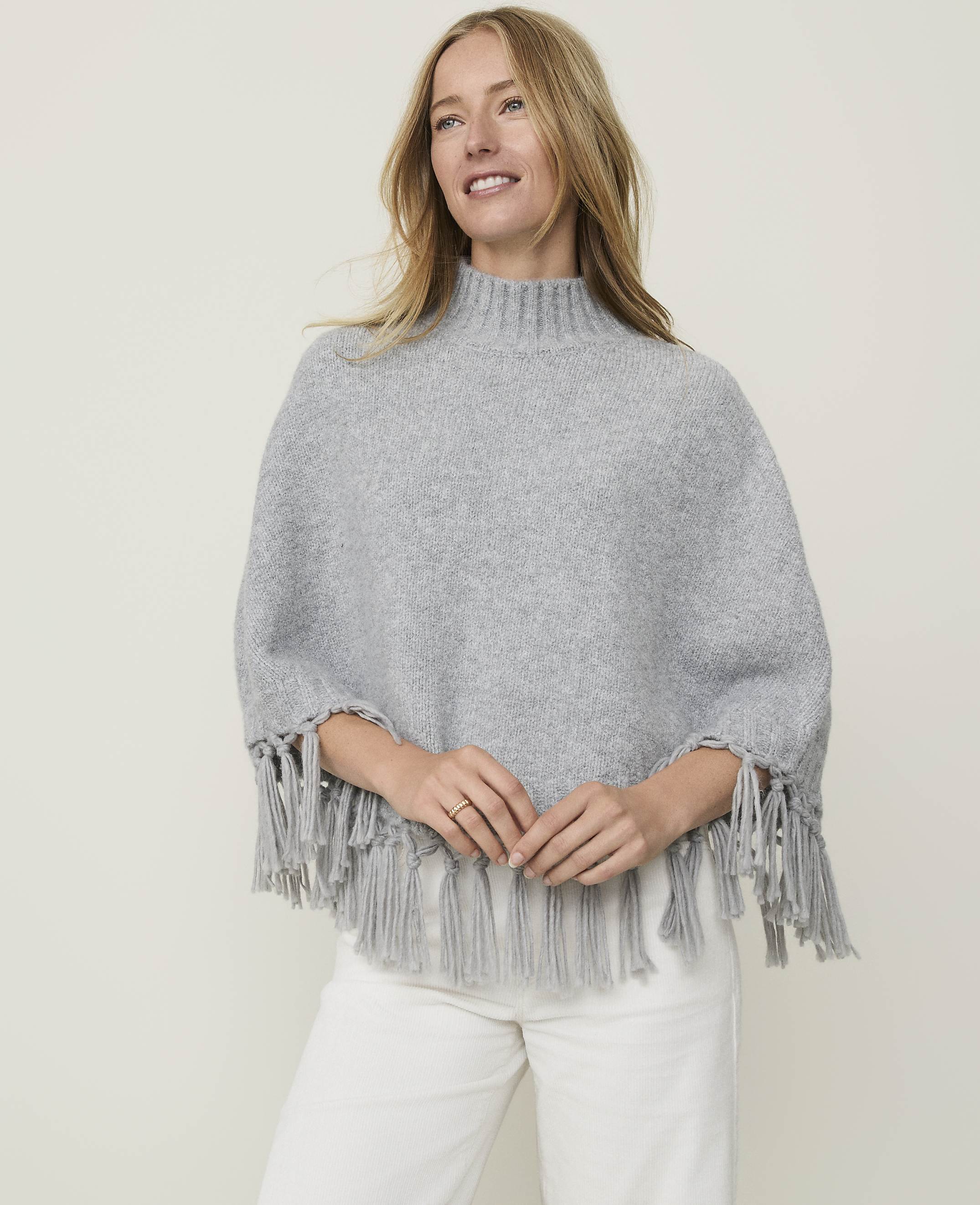 A blonde woman in a grey fringed poncho and white pants smiles against a plain, light grey background.