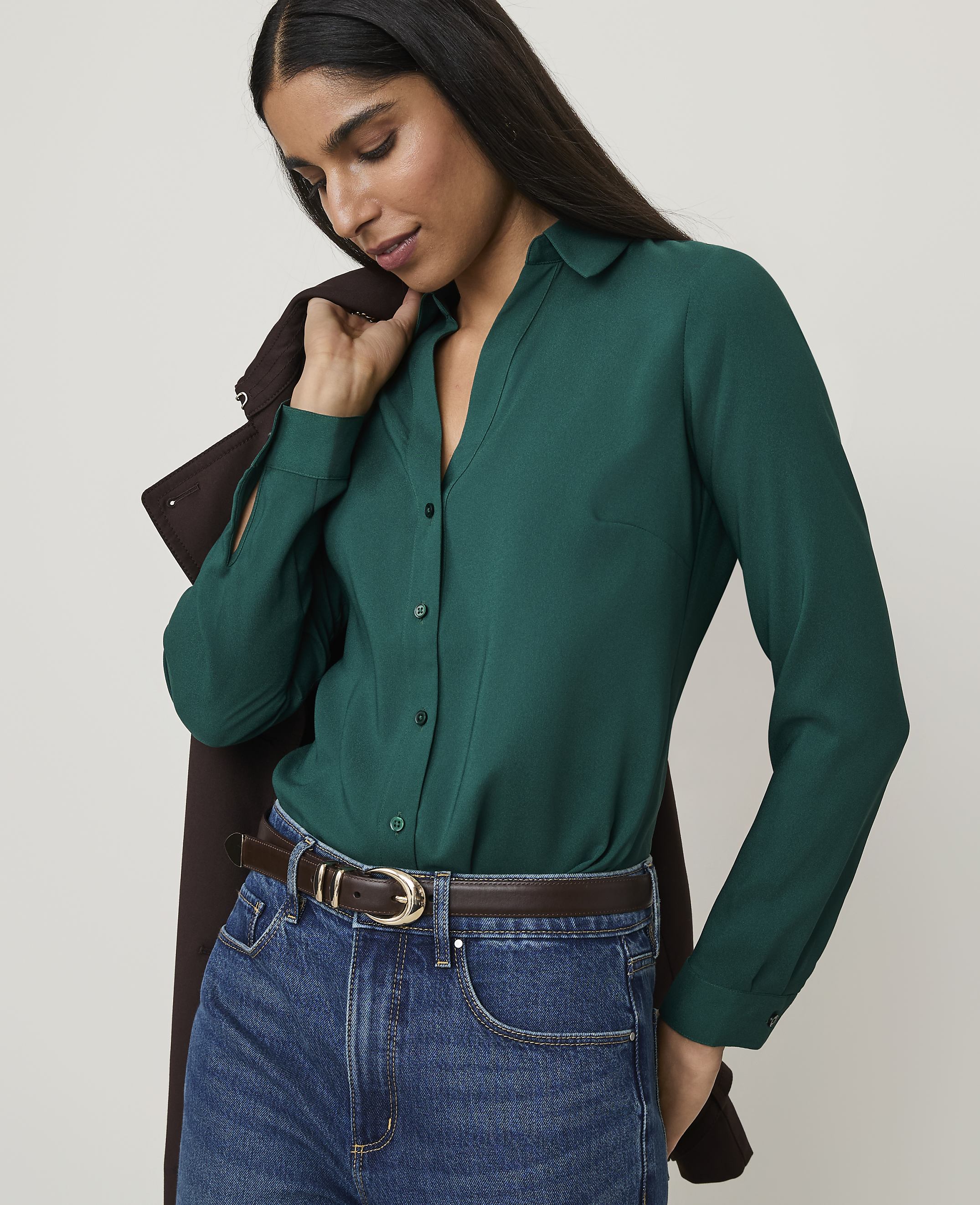 A woman in a green blouse and jeans stands against a plain background, with a brown jacket draped over her shoulder.