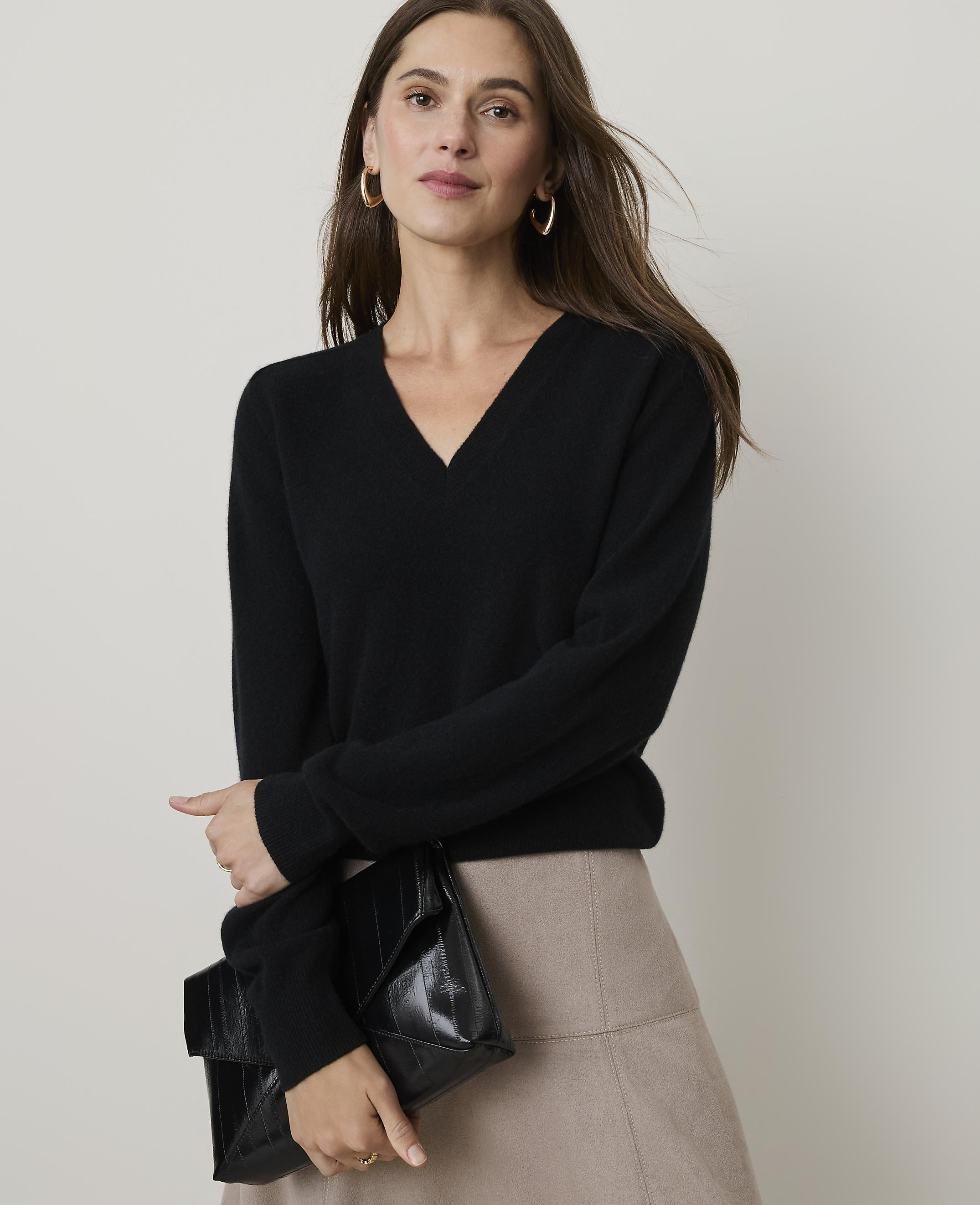 A woman in a black v-neck sweater and beige skirt holds a black clutch in front of a plain background.