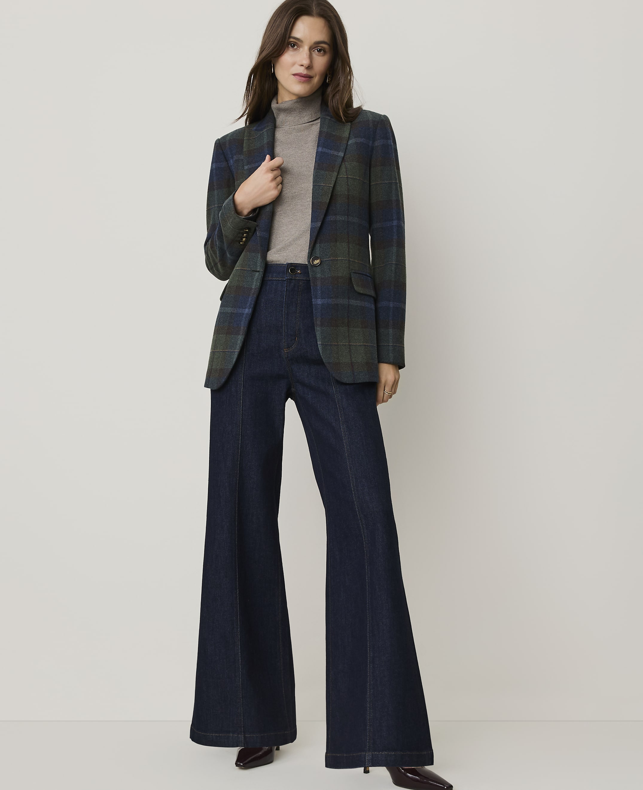 A woman in a plaid blazer and wide-leg jeans stands against a plain, light-colored background.