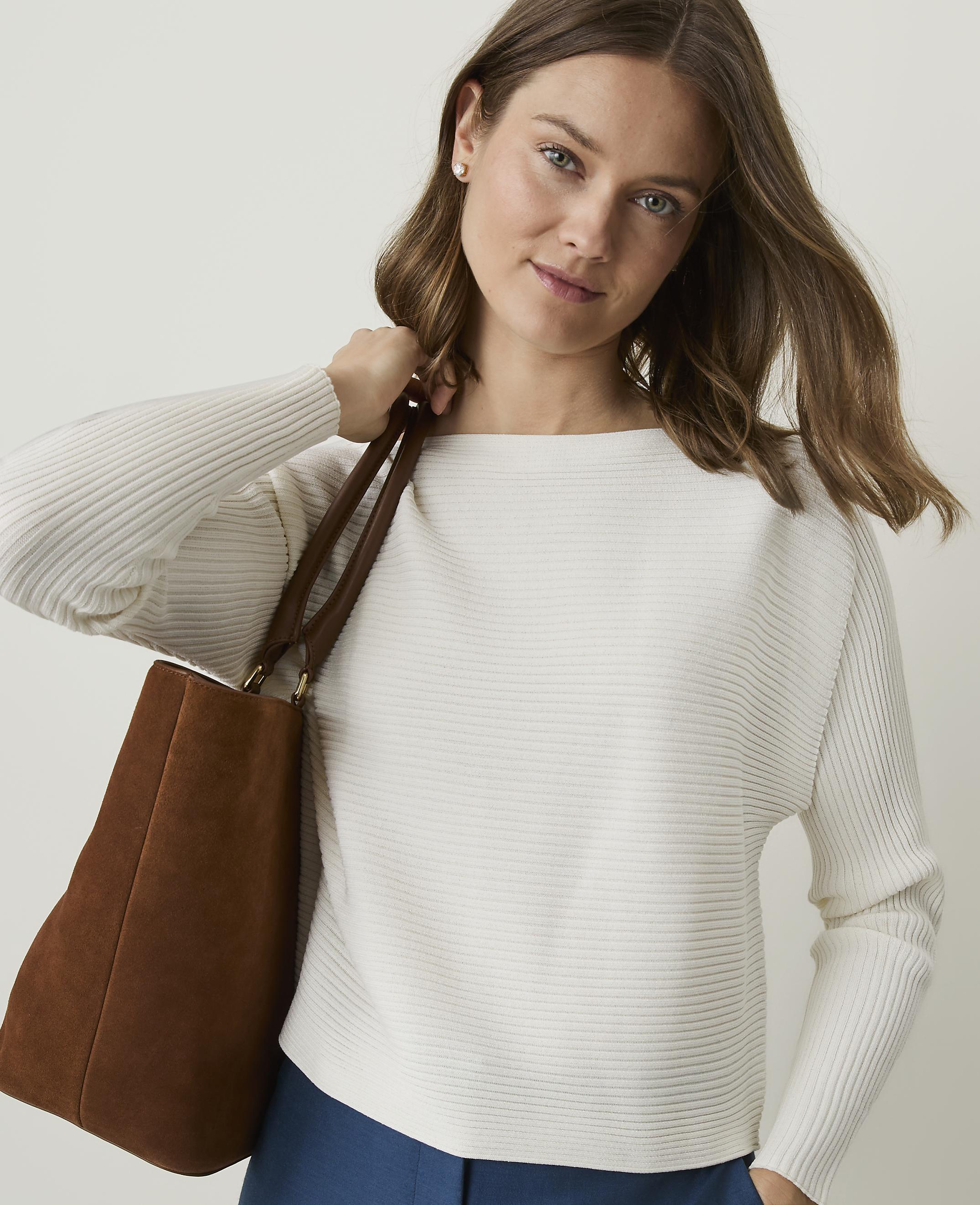 A woman in a white ribbed sweater and blue pants carries a brown tote bag against a plain background.