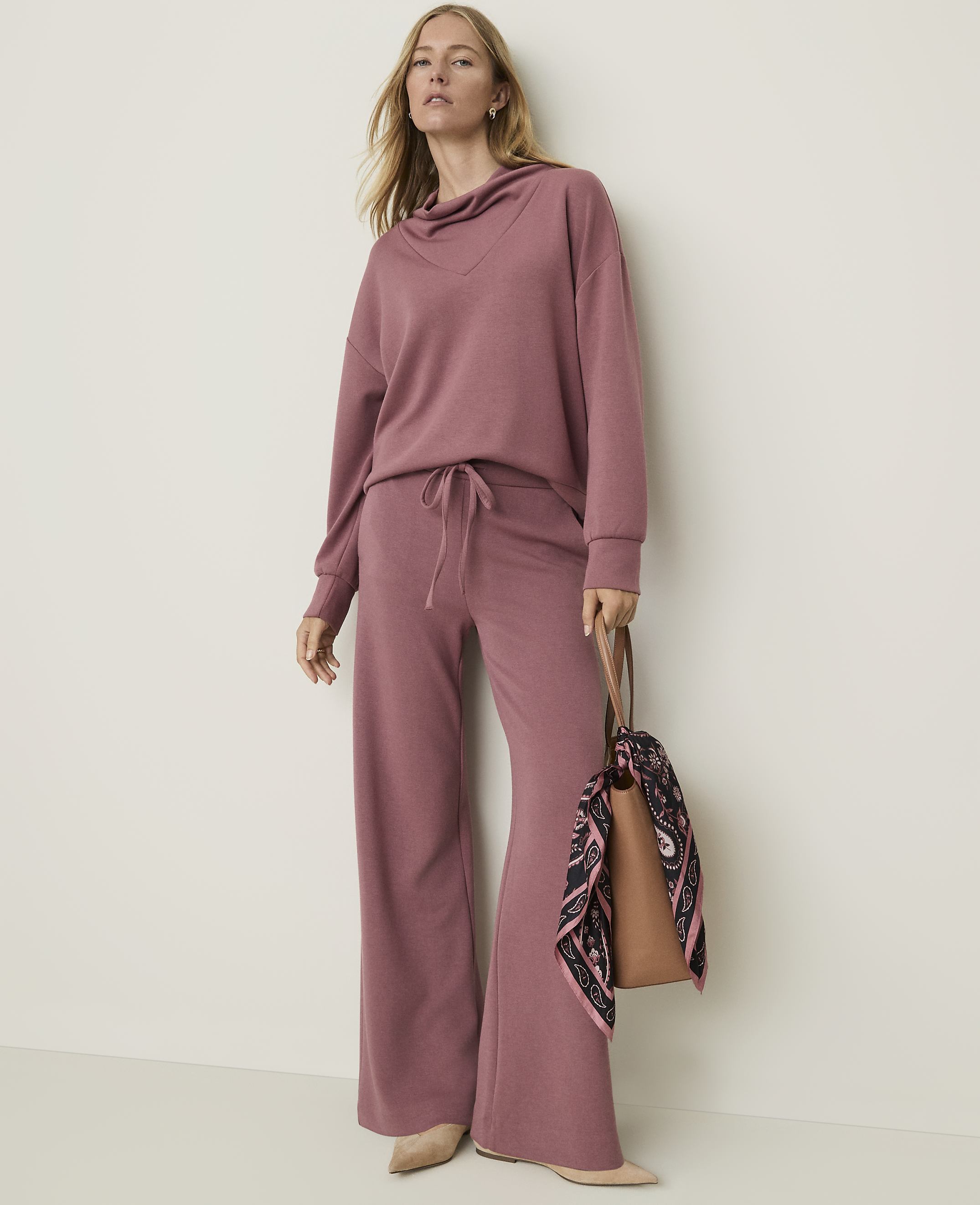 A woman in a mauve loungewear set stands against a plain, light-colored wall, holding a brown tote bag with a patterned scarf.