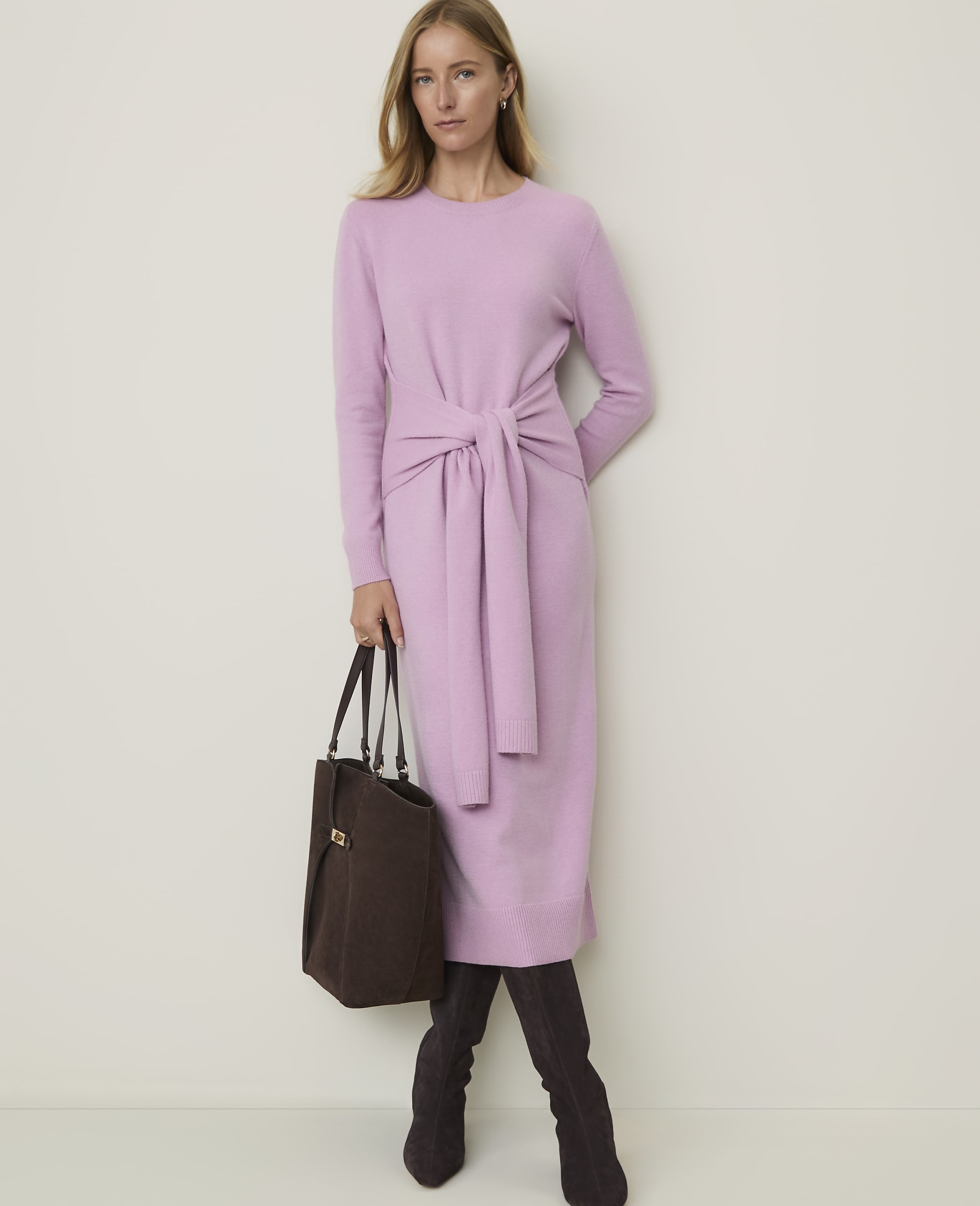 A woman in a light purple dress and dark boots stands with a brown bag against a plain white background.