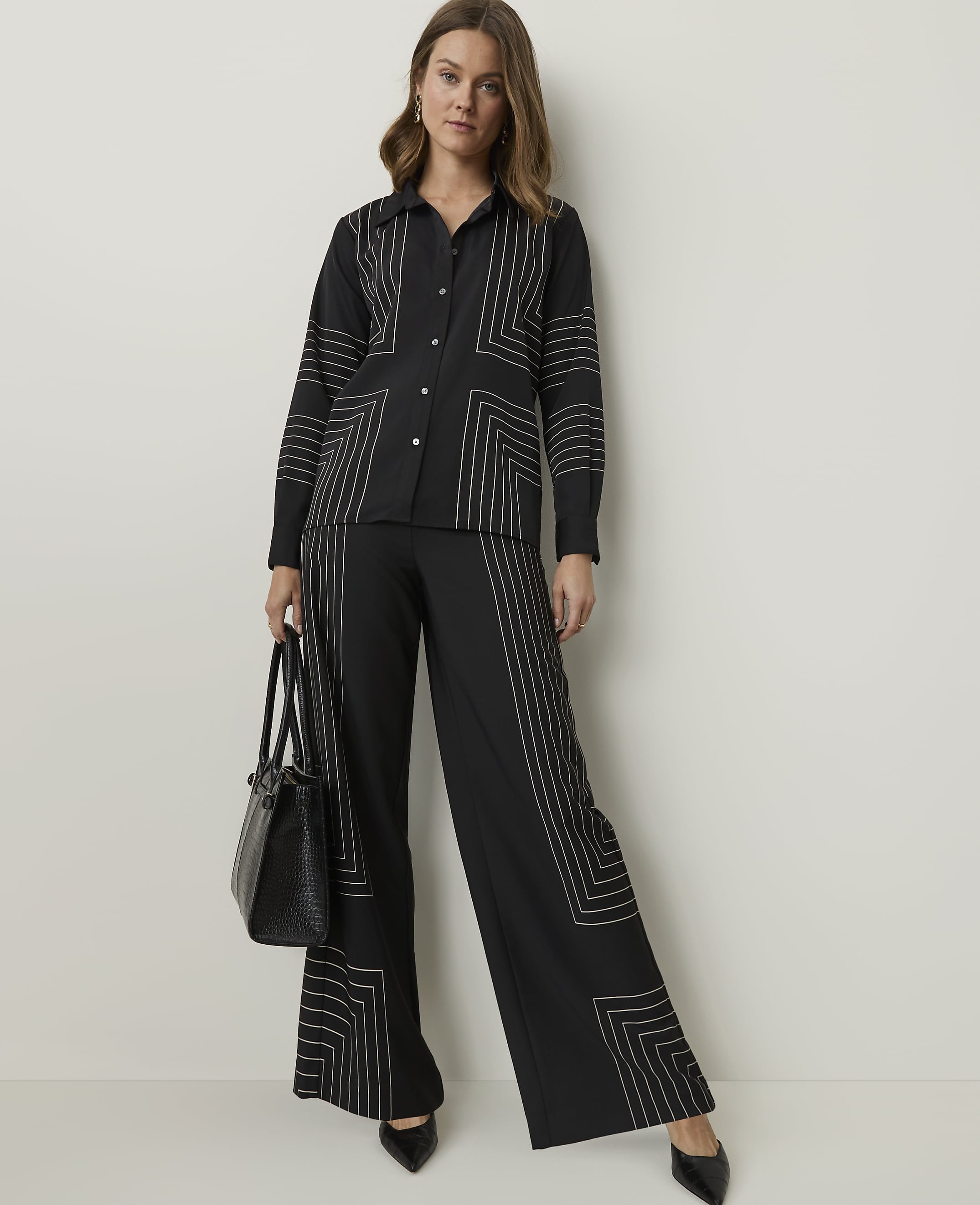 A woman in a black patterned shirt and wide-leg pants stands against a plain white background, holding a black handbag.
