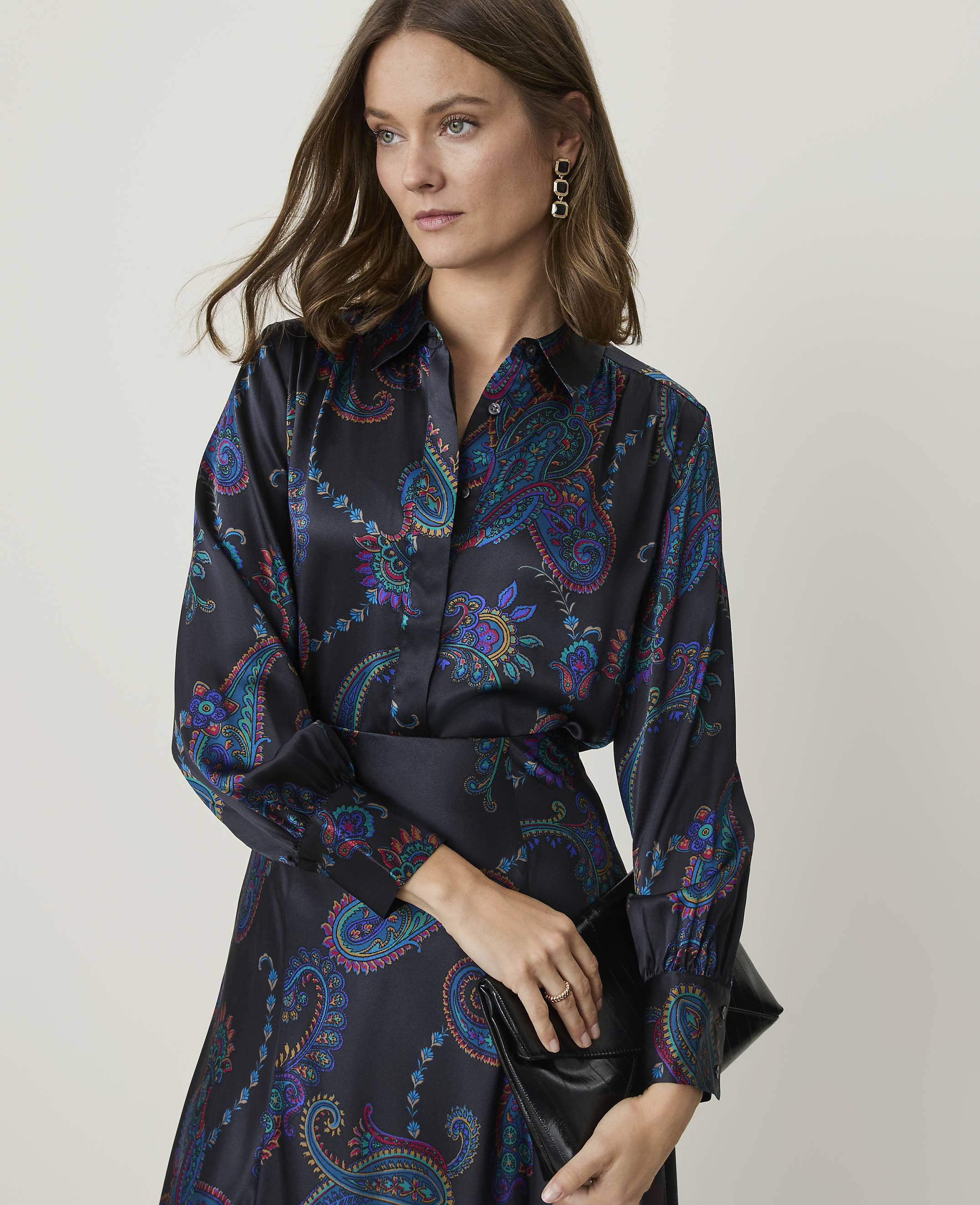 A woman in a dark, paisley-patterned blouse and skirt holds a black clutch against a plain, light background.
