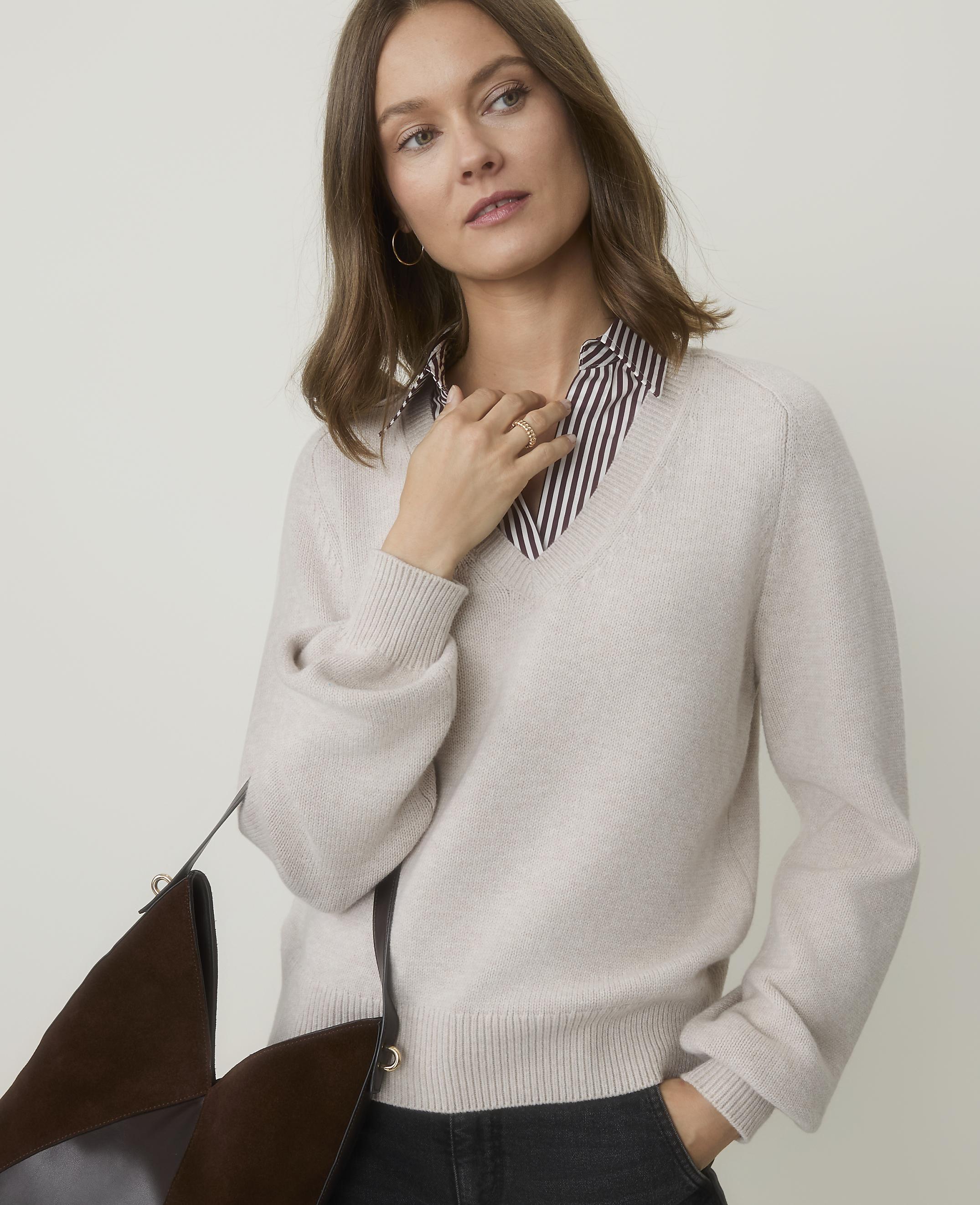 A woman in a cream v-neck sweater over a striped shirt stands against a plain, light background, with a brown bag in the foreground.
