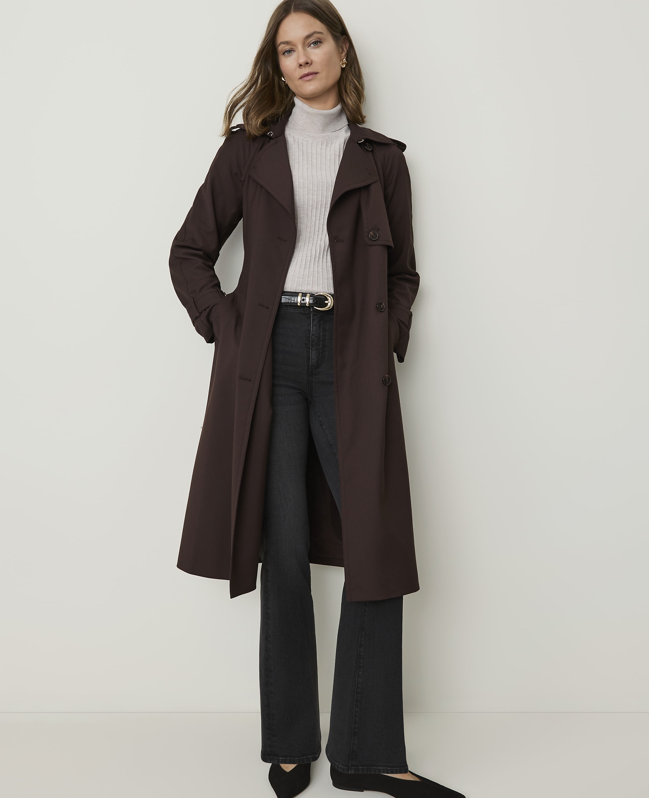 A woman in a brown trench coat and black flared jeans stands against a plain white background.
