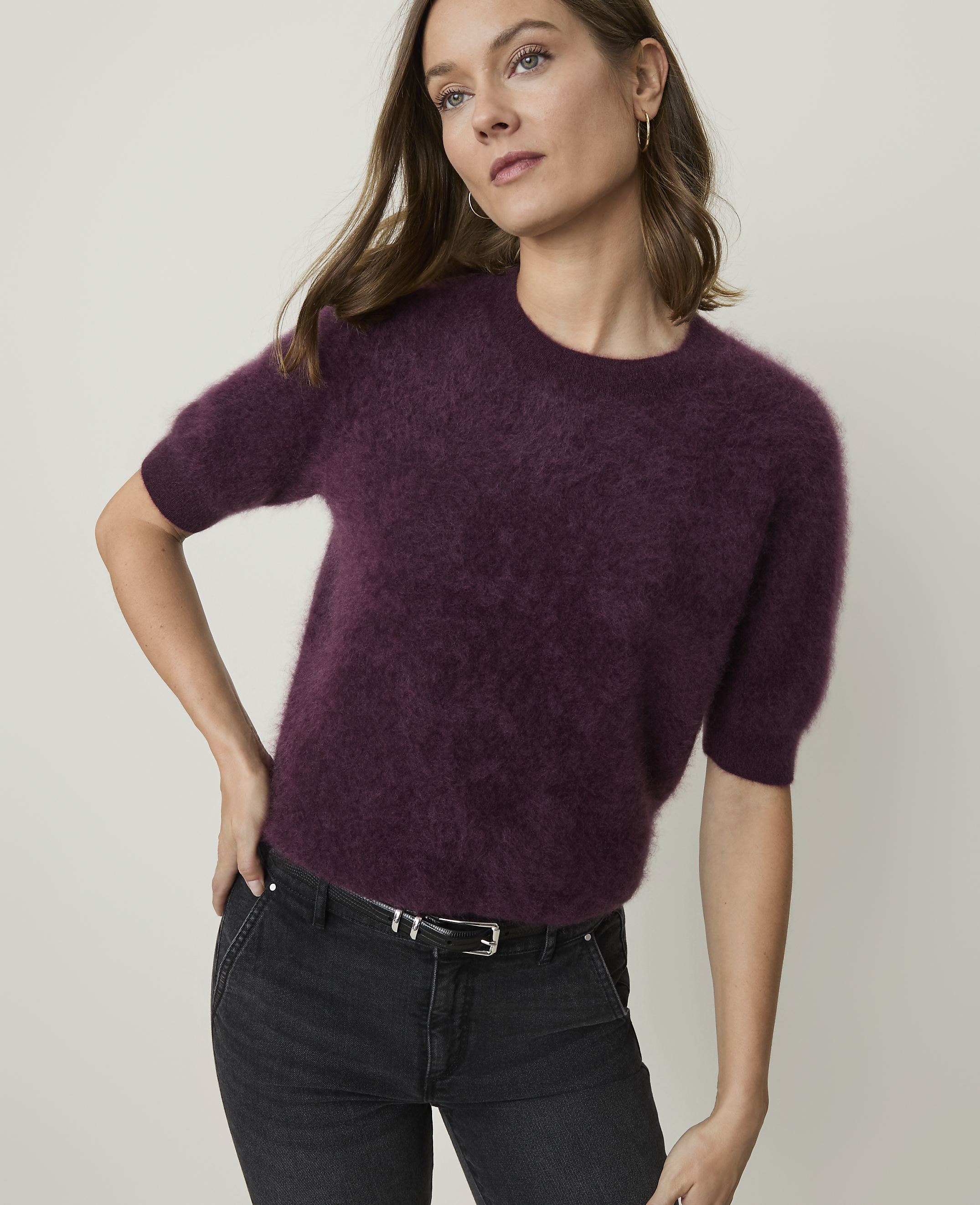 A woman in a fuzzy plum-colored sweater and dark jeans stands against a plain, light gray background.