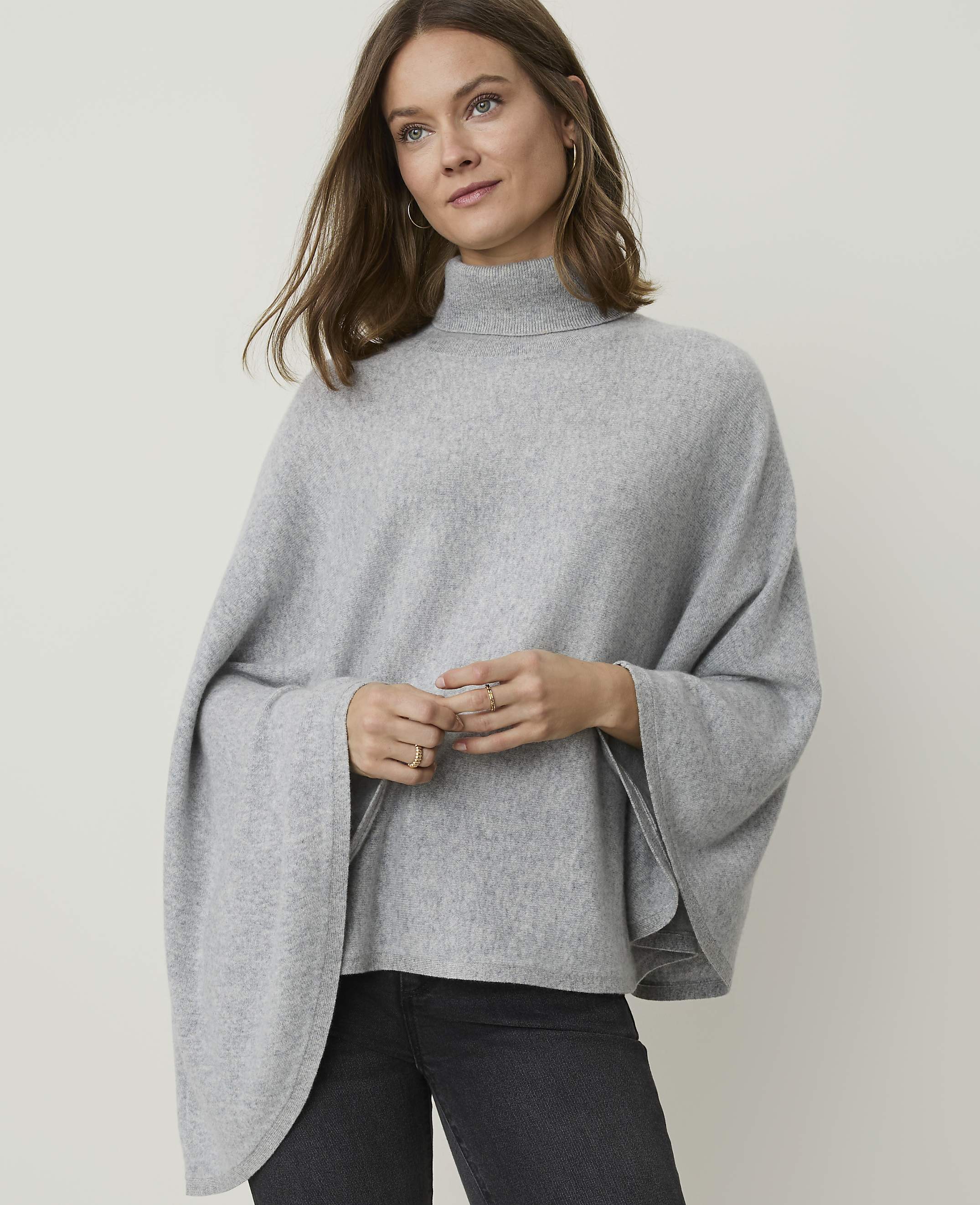 A woman in a grey turtleneck poncho stands against a plain grey background.