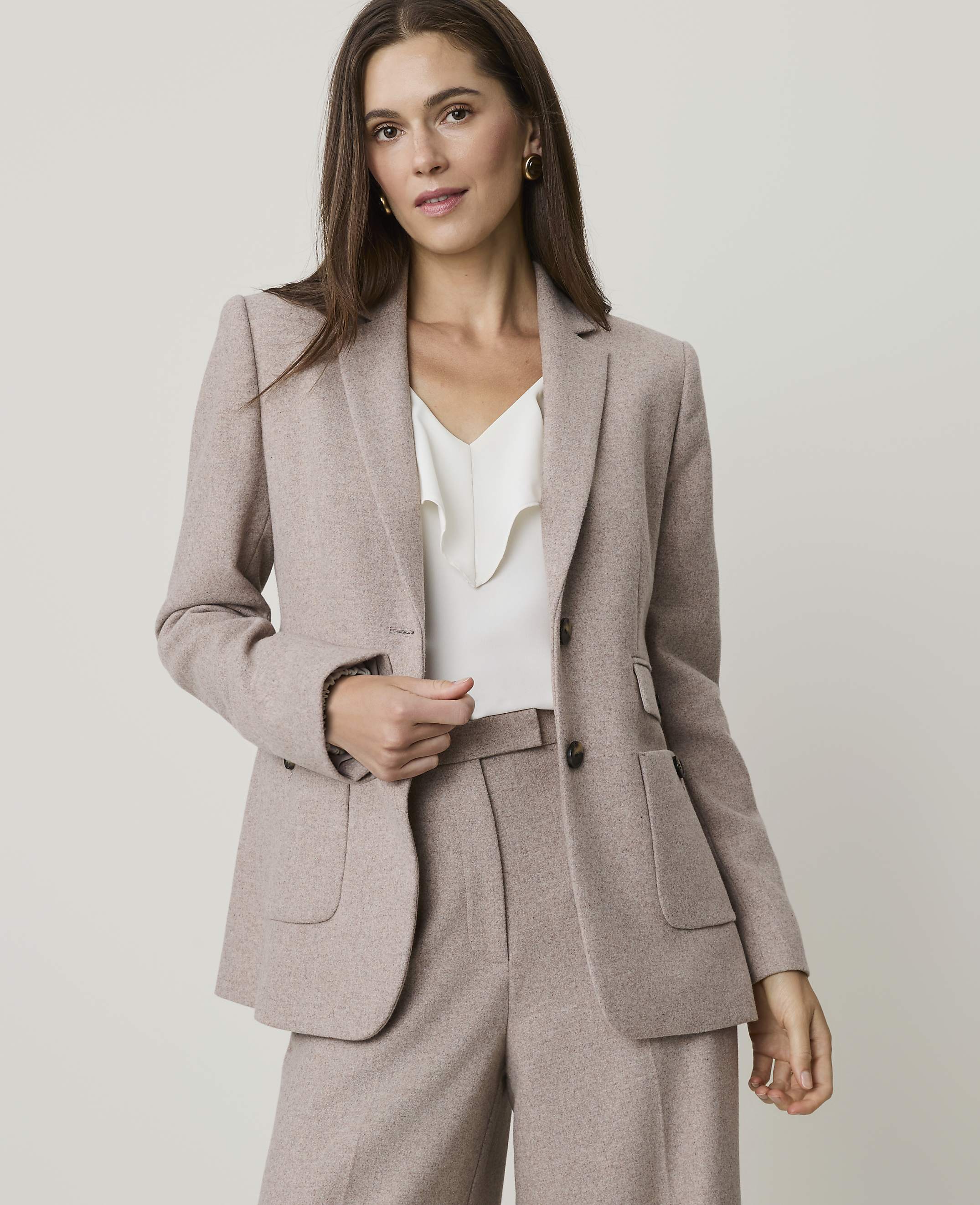 A woman in a beige blazer and pants stands against a plain gray background.