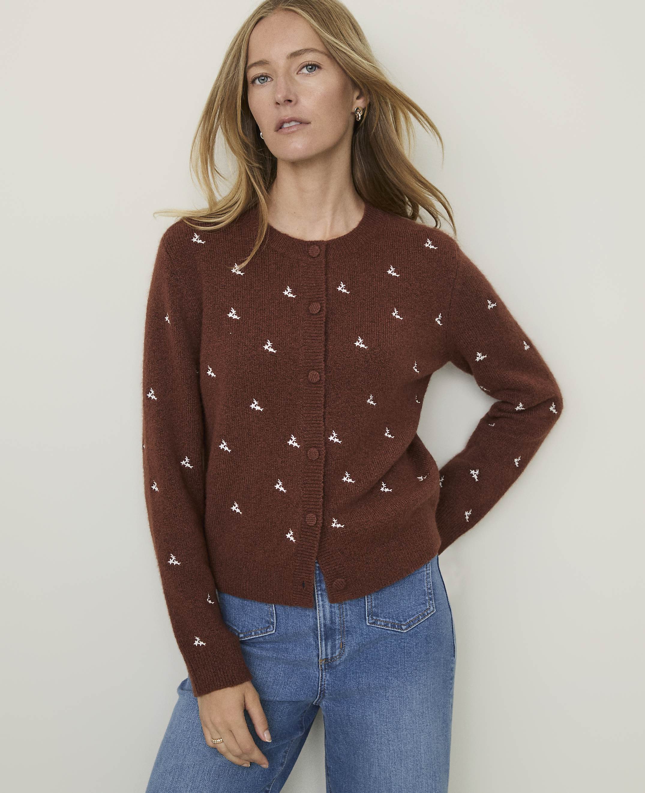 A woman in a brown embroidered cardigan and blue jeans stands against a plain, light-colored background.