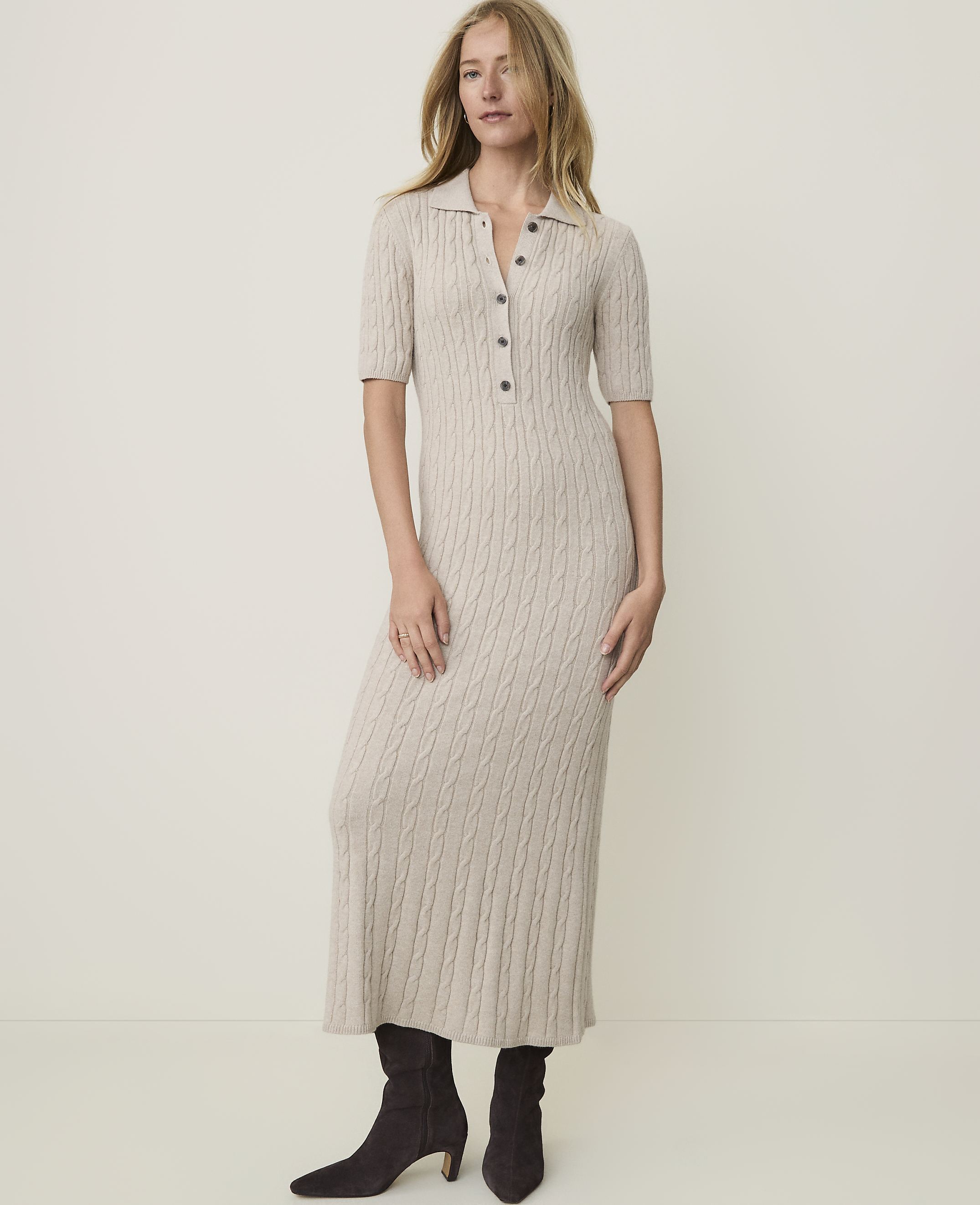 A woman in a beige cable-knit midi dress and dark boots stands against a plain, light-colored background.