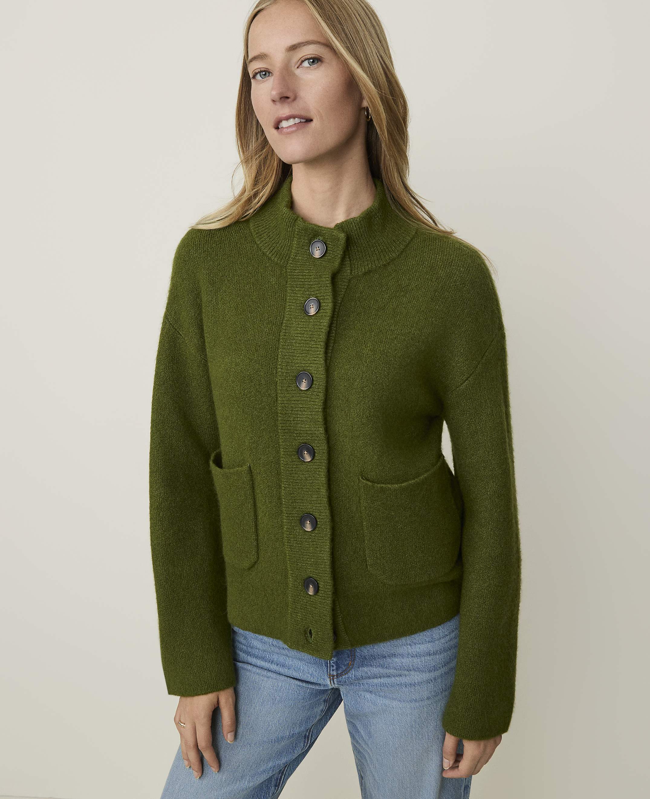 A woman in a green cardigan and jeans stands against a plain, light background.