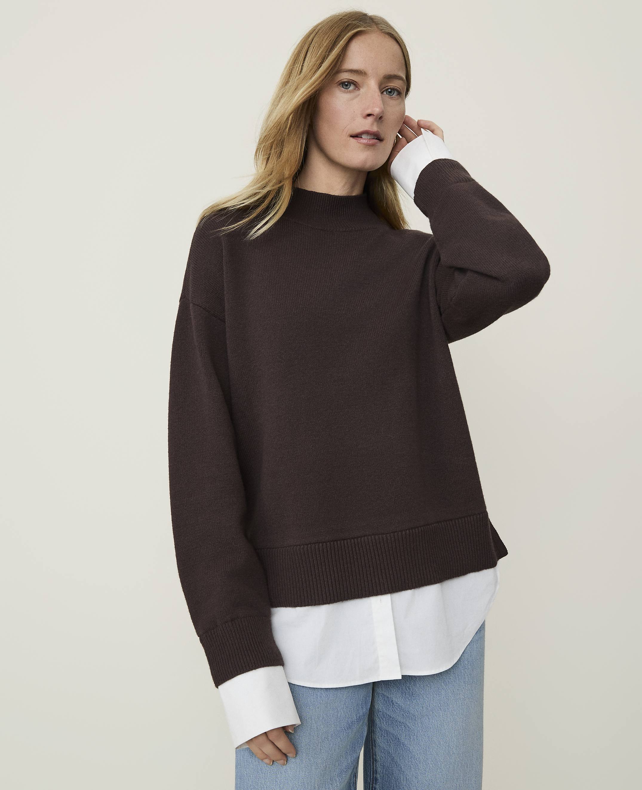 A woman in a brown sweater layered over a white shirt stands against a plain, light-colored background.