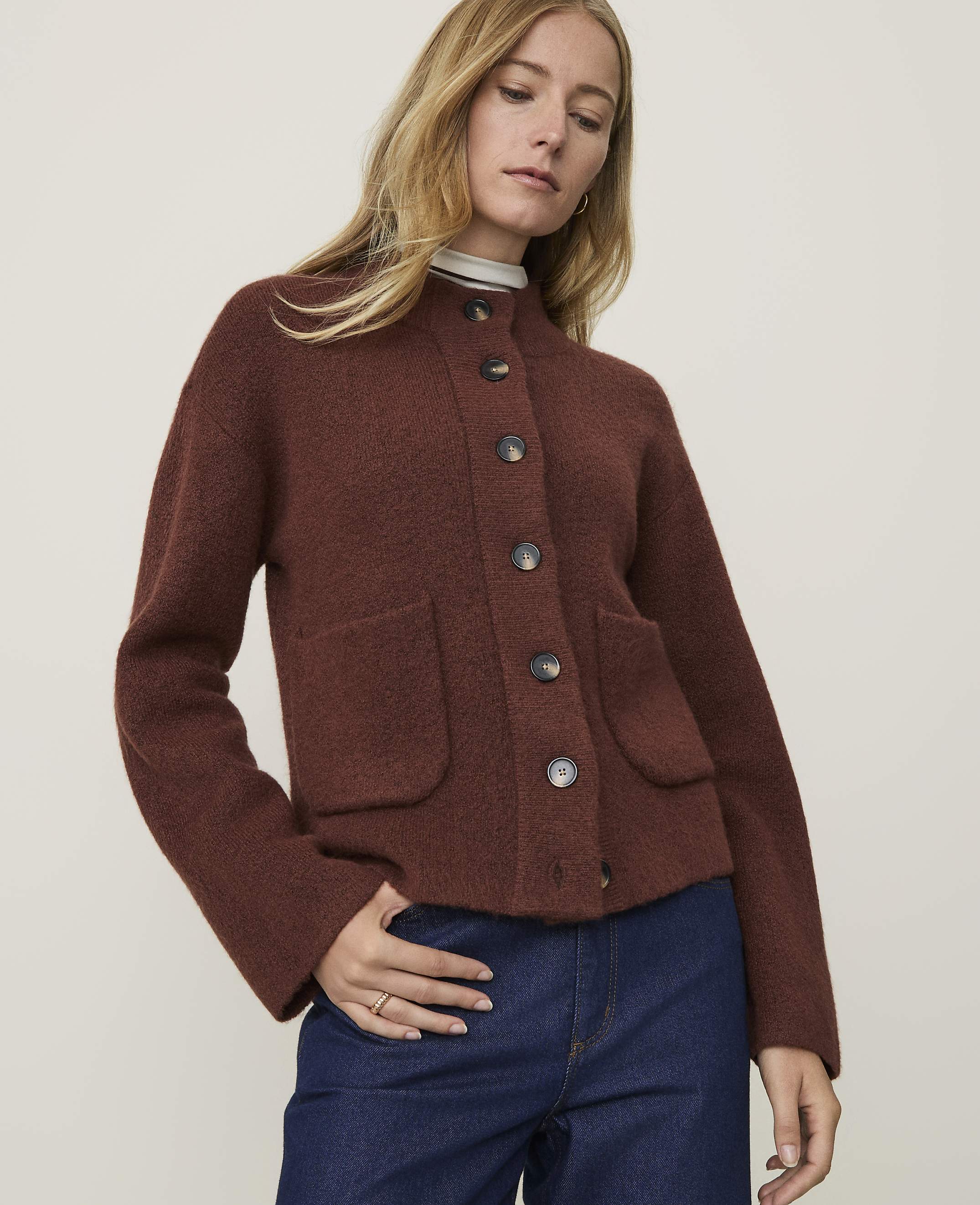 A woman in a brown cardigan and blue jeans stands against a plain, light-colored background.